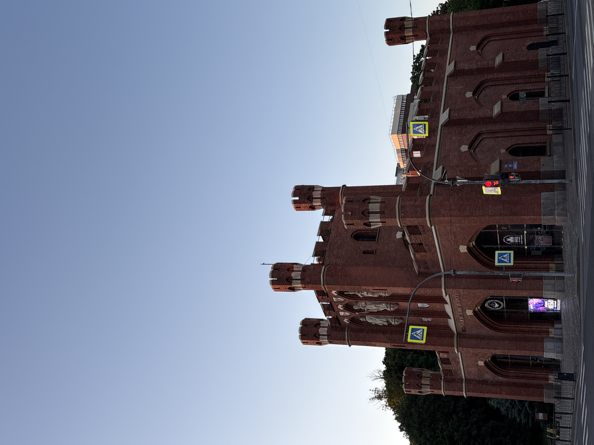 The image captures the Royal Gate, a historic landmark in Kaliningrad, Russia, bathed in the warm glow of late afternoon or early morning sunlight. The gate, constructed from red brick, features a formidable, castle-like design with multiple turrets and arched entrances. Statues adorn the facade above the central archway. Pedestrian crossing signs, a traffic light with a red signal, and street markings are visible, indicating the gate's location at a street intersection. Trees and foliage are present in the background, with a clear, pale blue sky overhead. The scene appears to be a static street view, with no people clearly visible in the foreground.