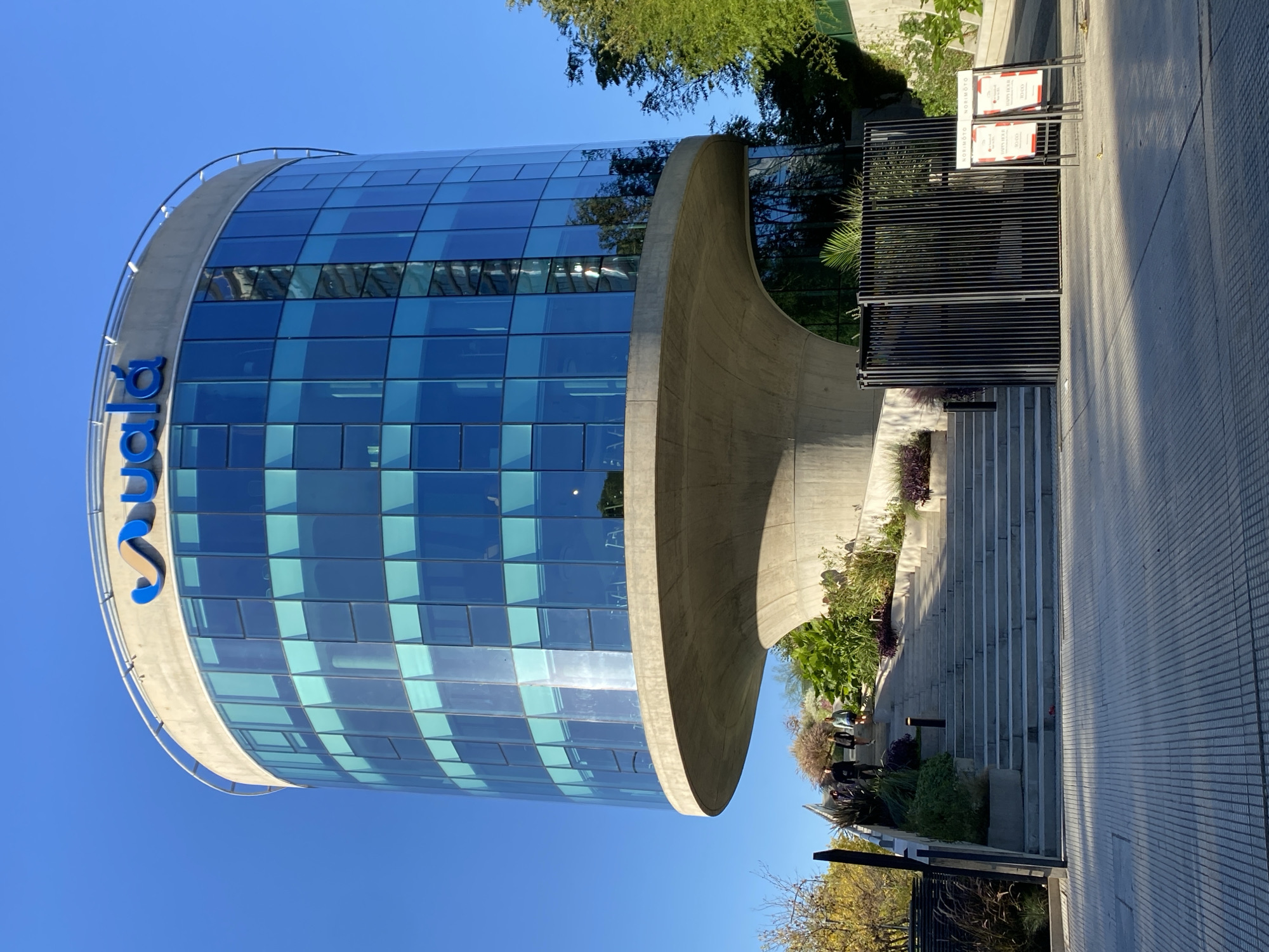 The image captures the Suela building in Buenos Aires, Argentina, showcasing a modern architectural design with a cylindrical glass tower and a prominent concrete support structure. The building features a grid-like pattern on its glass facade and the word "Suela" in blue letters on its upper portion. Adjacent to the building is a staircase leading up, bordered by greenery and decorative plants. A dark metal railing stands near the entrance, with signs attached. The surrounding area appears to be a paved walkway. The sky is clear and blue, suggesting a sunny day.
