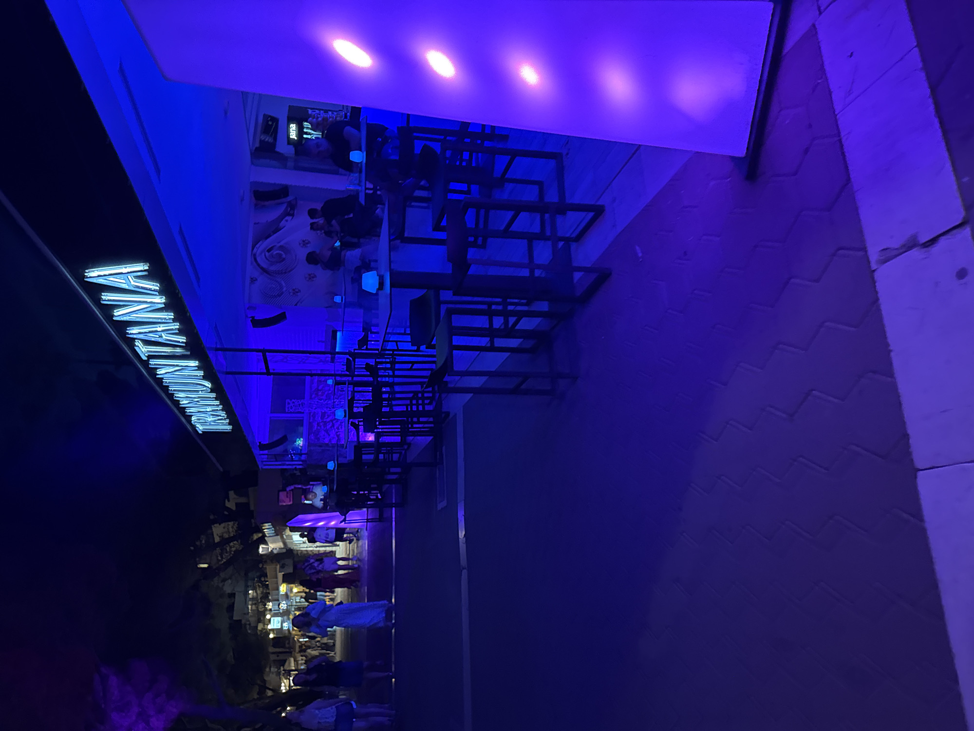 The image depicts a nighttime scene on a promenade in Makarska, Croatia, likely a commercial area or waterfront. A prominent neon sign, reading "TRAMUNTANA," is illuminated in blue above a building that appears to be a bar or restaurant. The establishment features outdoor seating with high-top tables and stools, illuminated by blue lights. Several individuals are visible on the promenade, some walking and others seated at the tables. The ground is paved with textured tiles, and there is a large, illuminated column or structure to the right of the frame emitting a soft purple glow. The overall atmosphere is one of a lively evening in a tourist destination.