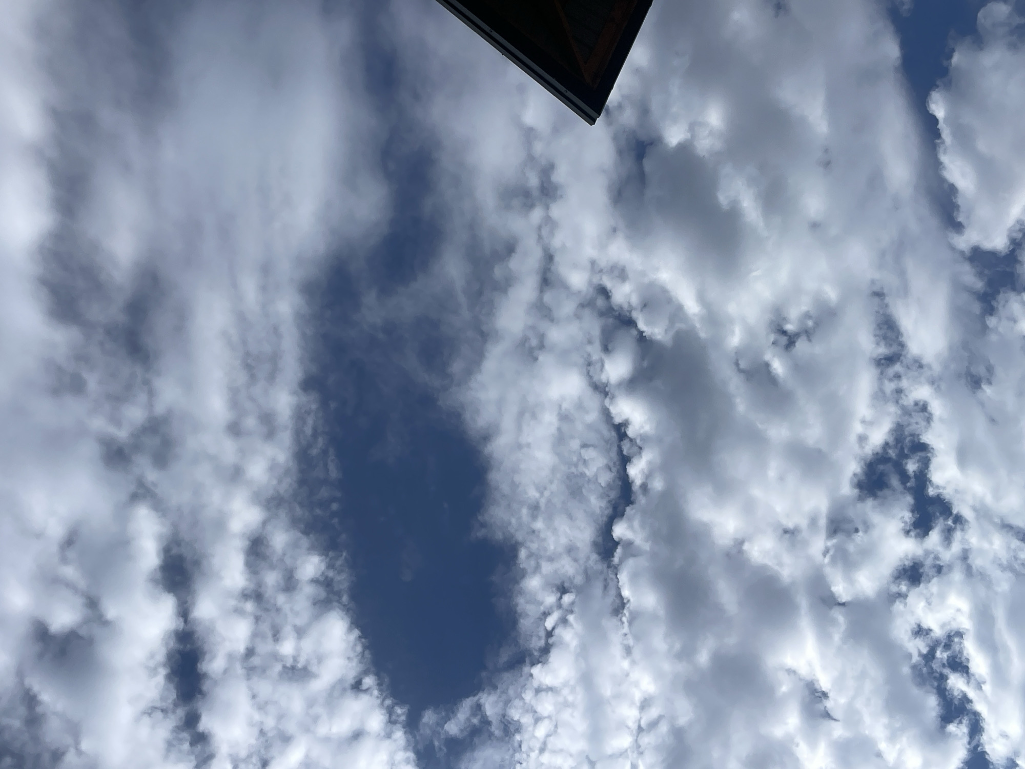 The image shows a close-up view of a blue sky filled with a mix of white and gray clouds. The clouds appear fluffy and scattered, with some areas showing darker, more ominous shades. In the upper right corner, a portion of a dark-colored roofline with visible wooden beams is visible, suggesting the perspective is from below, looking up. The environment appears to be outdoors, possibly on a partly cloudy day. Without additional context or landmarks, it is not possible to determine the specific location in Tashkent, Uzbekistan.