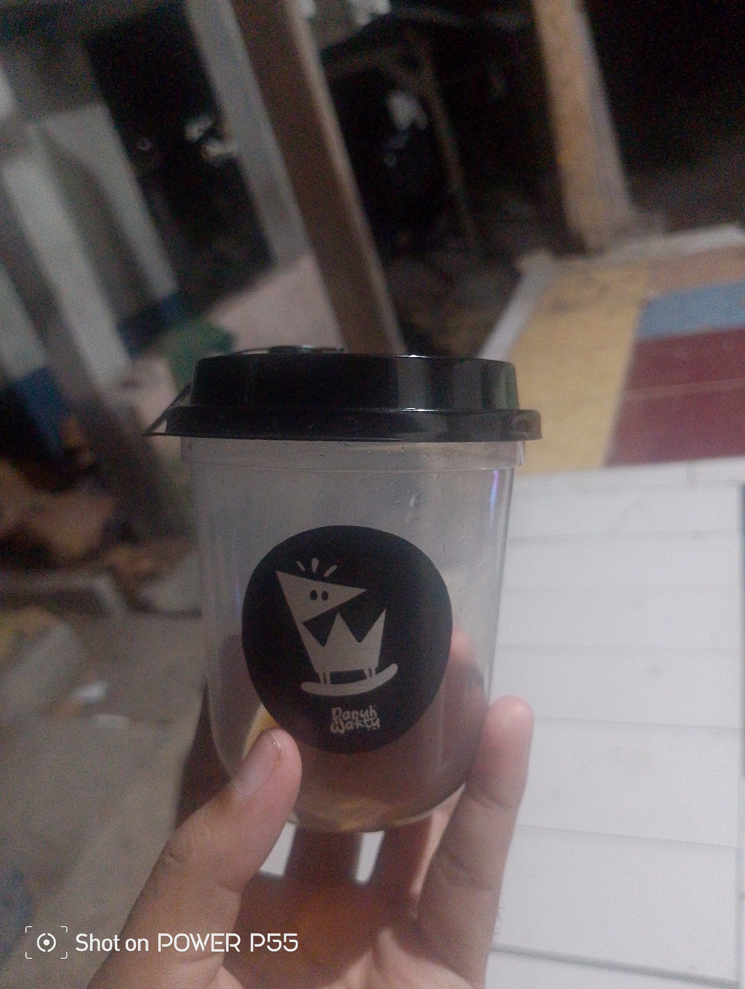 A close-up, dimly lit, indoor shot shows a hand holding a clear plastic cup with a black lid. A circular black sticker with a white logo of a crown with two dots and radiating lines is affixed to the cup, along with the text "Daruh Waktu". The background is blurred, showing vertical wooden beams and hints of colored panels. The bottom left corner of the image displays "Shot on POWER P55". No specific location in Yamansari, Indonesia, can be determined from the image.
