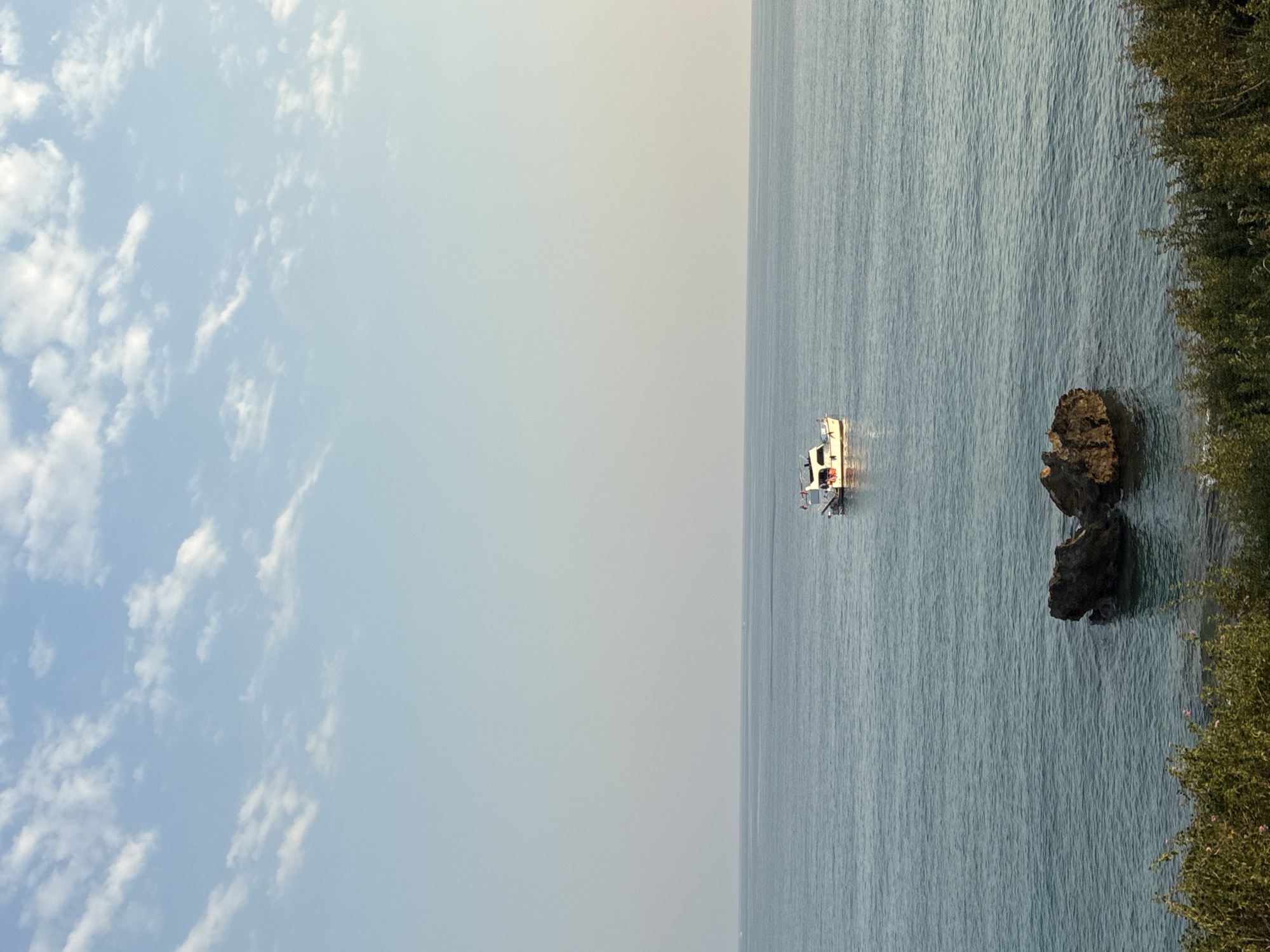 A motor yacht is at anchor in the Mediterranean Sea, approximately 50 meters from the coast of Alanya, Turkey. The yacht is white with a red stripe along its waterline. It has an enclosed cabin and an open deck at the stern. Two people are visible on the deck of the yacht. In the foreground, two large rocks are partially submerged in the water, with lush green vegetation growing on the shoreline behind them. The sky is a pale blue with scattered white clouds.