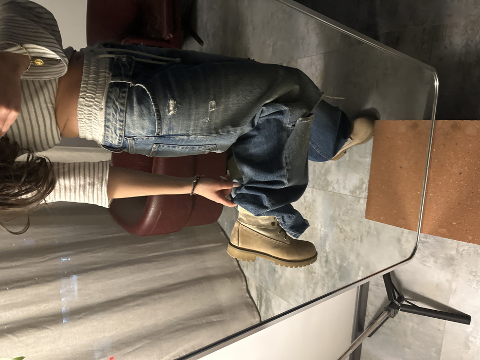 An indoor scene in Yerevan, Armenia, depicts a partially visible individual in front of a mirror, capturing their reflection. The subject, appearing to be a female, wears a light-colored, horizontally striped crop top exposing the midriff, high-waisted, distressed light-wash blue jeans with an elastic waistband and drawstring, and tan-colored lace-up boots with thick, ridged soles. A gold-colored pendant necklace is visible on the subject's right chest, and a dark bracelet is on the left wrist.
The individual is standing, with the right leg lifted, showcasing the tan boot. The left hand is holding the cuff of the right pant leg, which is baggy and gathered around the ankle. The right arm is bent, with the hand partially obscured, suggesting it is holding a device to capture the image.
The environment, visible both directly and in reflection, features a light gray patterned floor. In the background, behind the subject's reflection, a light-colored curtain covers a window, through which blurred lights from an urban setting are faintly visible. Two dark red armchairs are partially reflected to the right of the subject. A metal tripod stand is visible on the left side, supporting what appears to be the mirror. A rectangular, orange-brown, brick-like object is positioned on the floor in the foreground, partly beneath the mirror's lower edge.