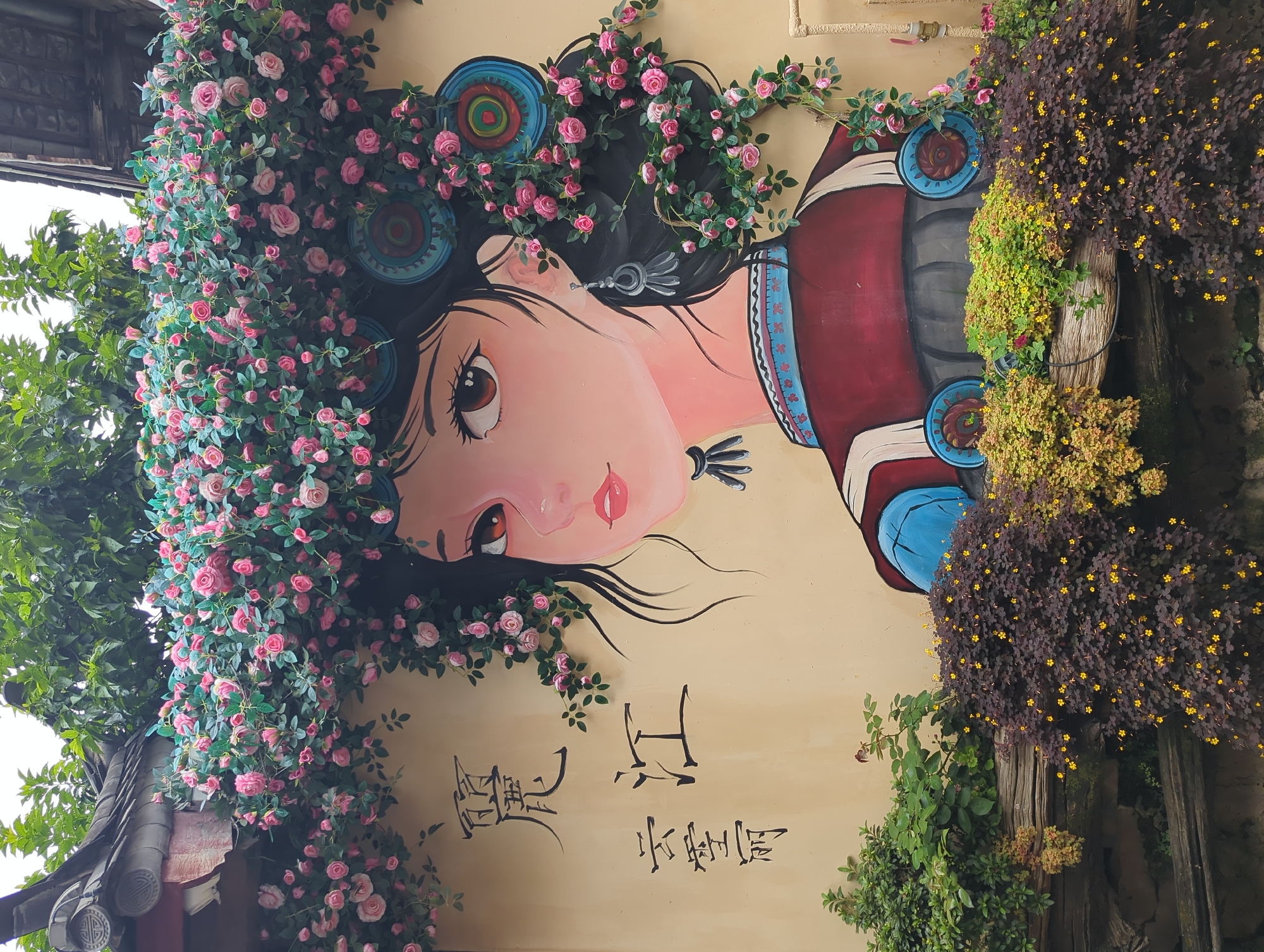 The image shows a mural of a woman on a tan-colored wall, located in Gucheng District, China. The mural depicts a woman with dark hair adorned with pink roses and green leaves. She has large, expressive brown eyes, pink lips, and wears a traditional outfit with blue and red accents. On the wall to the left of the mural, there are two lines of Chinese calligraphy. The mural is partially covered by an overhang with a dark, tiled roof and is decorated with trailing pink roses and green foliage that extends down the sides and over the woman's hair. In front of the wall, a low ledge is filled with various potted plants, including a dark leafy plant with small yellow flowers and a lighter green plant with yellow flowers. The overall scene suggests an outdoor decorative element, possibly in a courtyard or along a street in an area with traditional architecture.