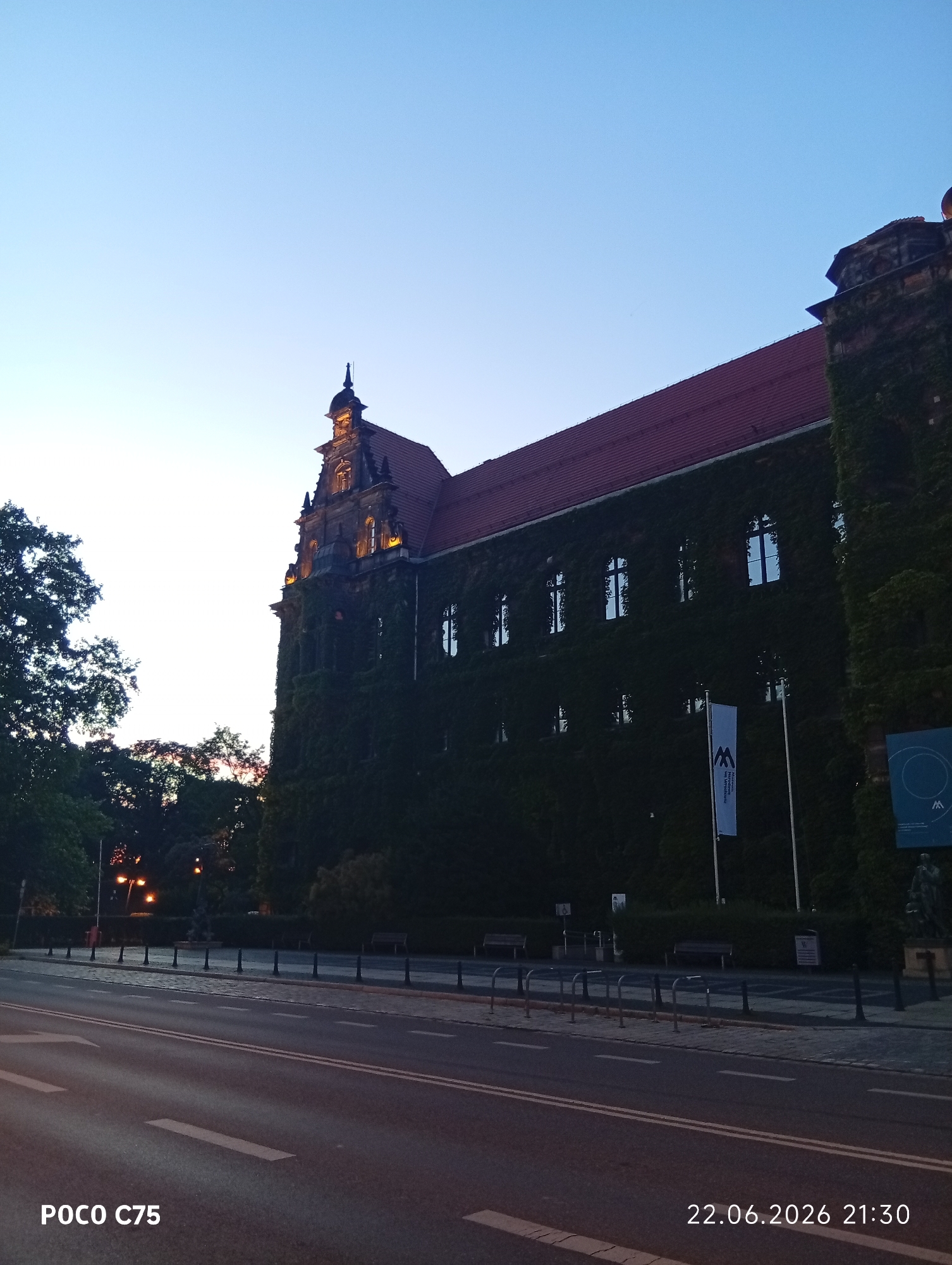 The image captures a building in Wrocław, Poland, at dusk on June 22, 2026, at 21:30. The building, likely the National Museum in Wrocław, is covered in ivy and features a red tiled roof. Several windows are visible on the building's facade. A white banner with a blue logo hangs on the building. In front of the building, there is a street with lane markings and a sidewalk lined with bollards and benches. Trees are visible on the left side of the image, and streetlights illuminate the area in the background. The sky is a clear gradient of light blue, indicating the transition from day to night. The foreground shows a portion of the street. The bottom left corner displays "POCO C75" and the bottom right corner displays the date and time.