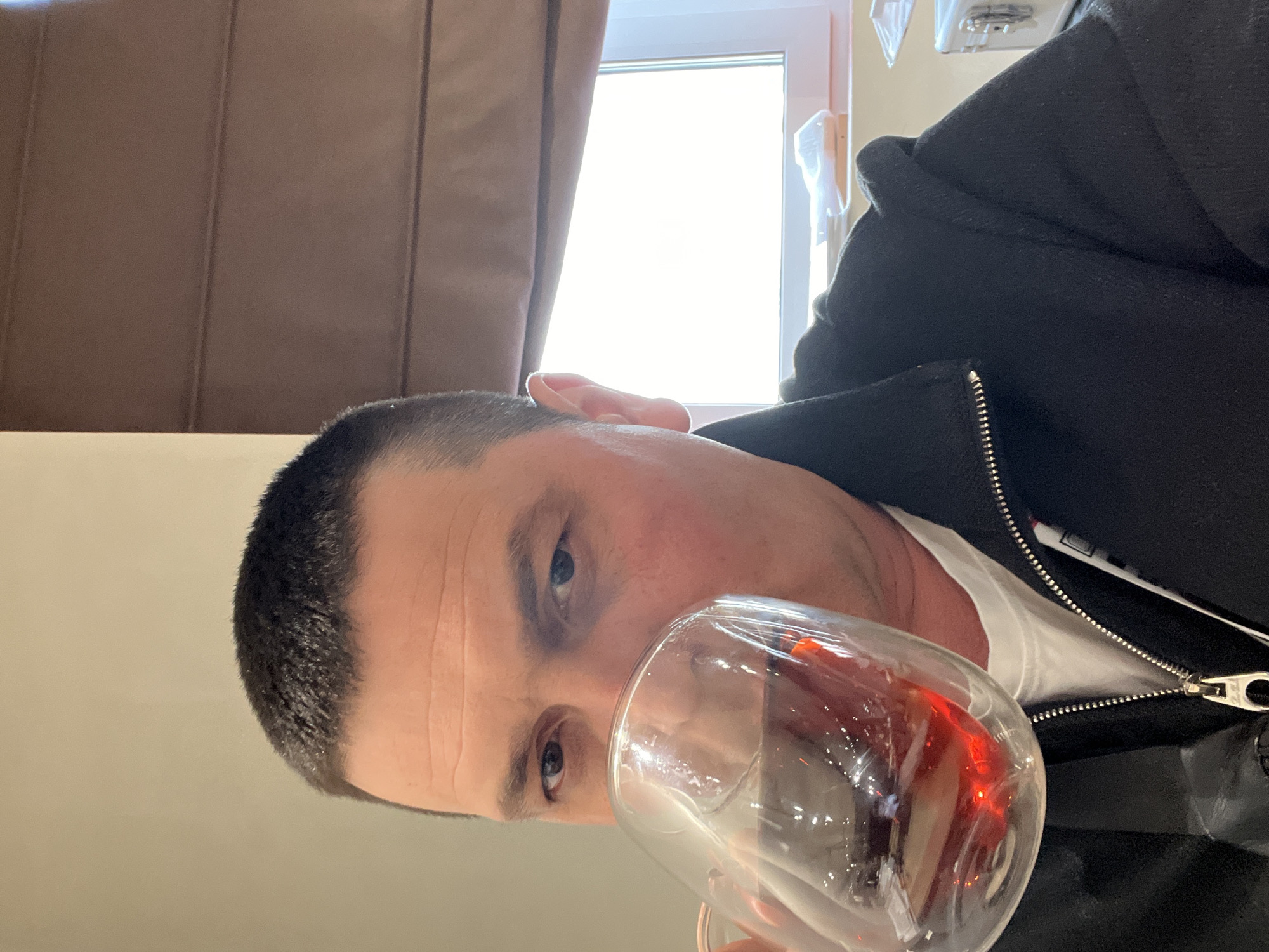 A man is shown from a close-up, canted perspective. He is fair-skinned with closely cropped dark hair and blue eyes. He is wearing a white undershirt and a black jacket with a zipper partially visible. The man is holding a clear glass containing a red liquid, possibly wine or juice, up to his face as if he is about to drink from it or is looking at the liquid. A window with bright light is visible in the background, partially obscured by a brown curtain. The setting appears to be indoors. The location and city are unknown.