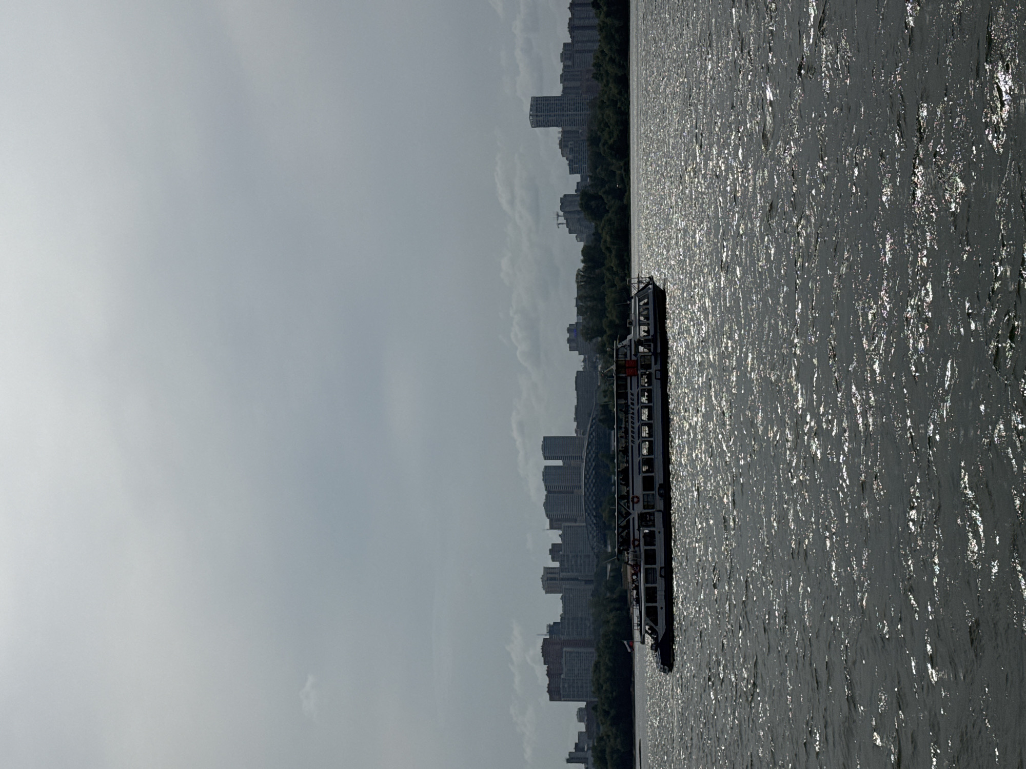 The image depicts a cloudy day on the Ob River in Novosibirsk, Russia. A white passenger boat with people on its decks sails across the water, its hull featuring the Cyrillic text "ПРokonьEcK". In the background, a dense line of trees separates the river from the city skyline, which is dominated by tall buildings of various architectural styles. A large, modern, dome-shaped structure is visible amongst the buildings. The sun reflects off the choppy surface of the river, creating a shimmering effect.