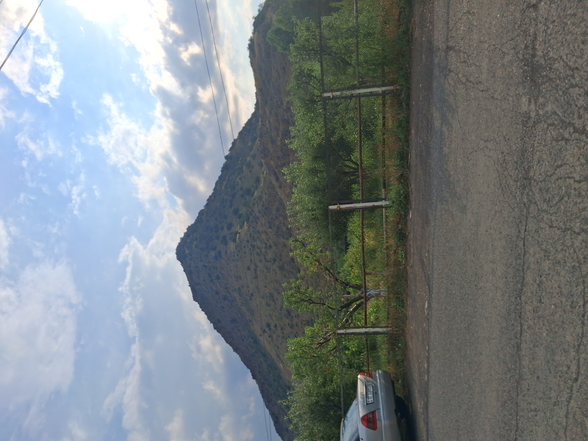 The image depicts a scene on the outskirts of Vayk, Armenia, featuring a prominent, cone-shaped mountain in the background under a partly cloudy sky. In the foreground, a silver Mercedes-Benz sedan is parked on the left side of a cracked asphalt road. A weathered metal fence runs horizontally across the middle ground, separating the road from a vegetated area that includes trees and bushes. The mountain is covered in sparse vegetation, giving it a dry, rugged appearance. The overall impression is a tranquil, rural landscape.