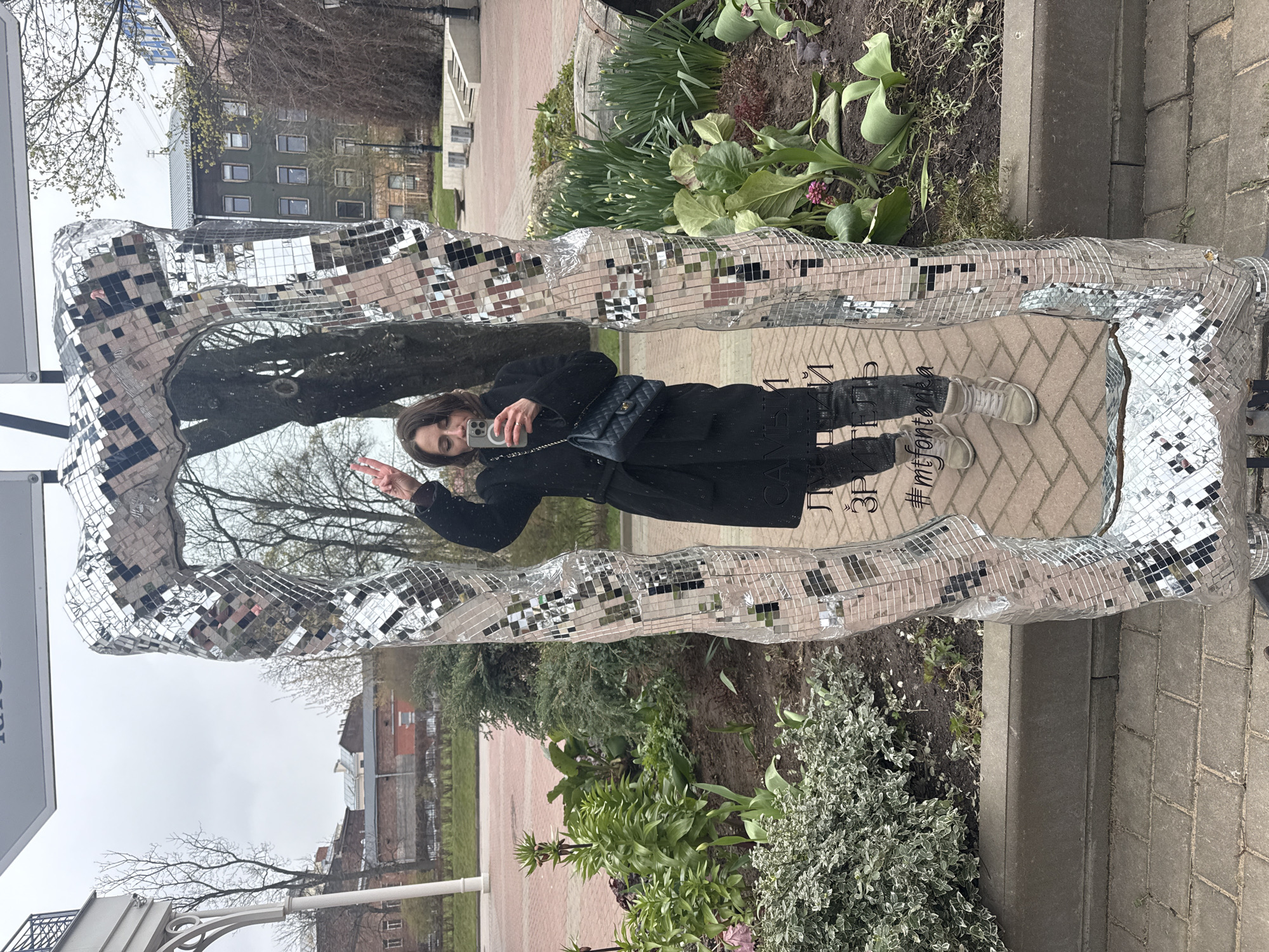 The image, when viewed in portrait orientation, captures a young woman reflected in a large, freestanding mirror frame. The frame is rectangular and entirely covered in small, square, reflective tiles, giving it a shimmering, disco ball-like appearance. The woman has long dark hair and is wearing a dark, possibly black, long coat, dark trousers, and light-colored sneakers. She carries a dark shoulder bag, which resembles a Chanel flap bag. She is holding a smartphone, seemingly taking a selfie or looking at the screen, and making a peace sign with her other hand, with a slight smile on her face.
The reflection in the mirror reveals the surrounding outdoor environment. In the background, beyond the woman, are trees – some still bare or just beginning to bud in early spring – and various buildings, including a multi-story structure with numerous windows and another lighter-colored building. The ground reflected appears to be a paved path or sidewalk.
The area around the mirror frame features garden beds with a variety of plants, including green leafy foliage, small buds, and shrubs, indicative of early spring. A brick-paved path and a concrete curb are also visible in the foreground. The overall scene suggests an urban park or public space on an overcast daytime.
Visible text, reflected on the paved surface near the woman's feet, includes Cyrillic letters spelling "Фонтанка" (Fontanka) and partially "#mtfontanka". Additional partial Cyrillic text, possibly "3P..." and parts of another word, can be seen near her shoulder. The mention of "Fontanka" strongly indicates the location as Saint Petersburg, Russia.