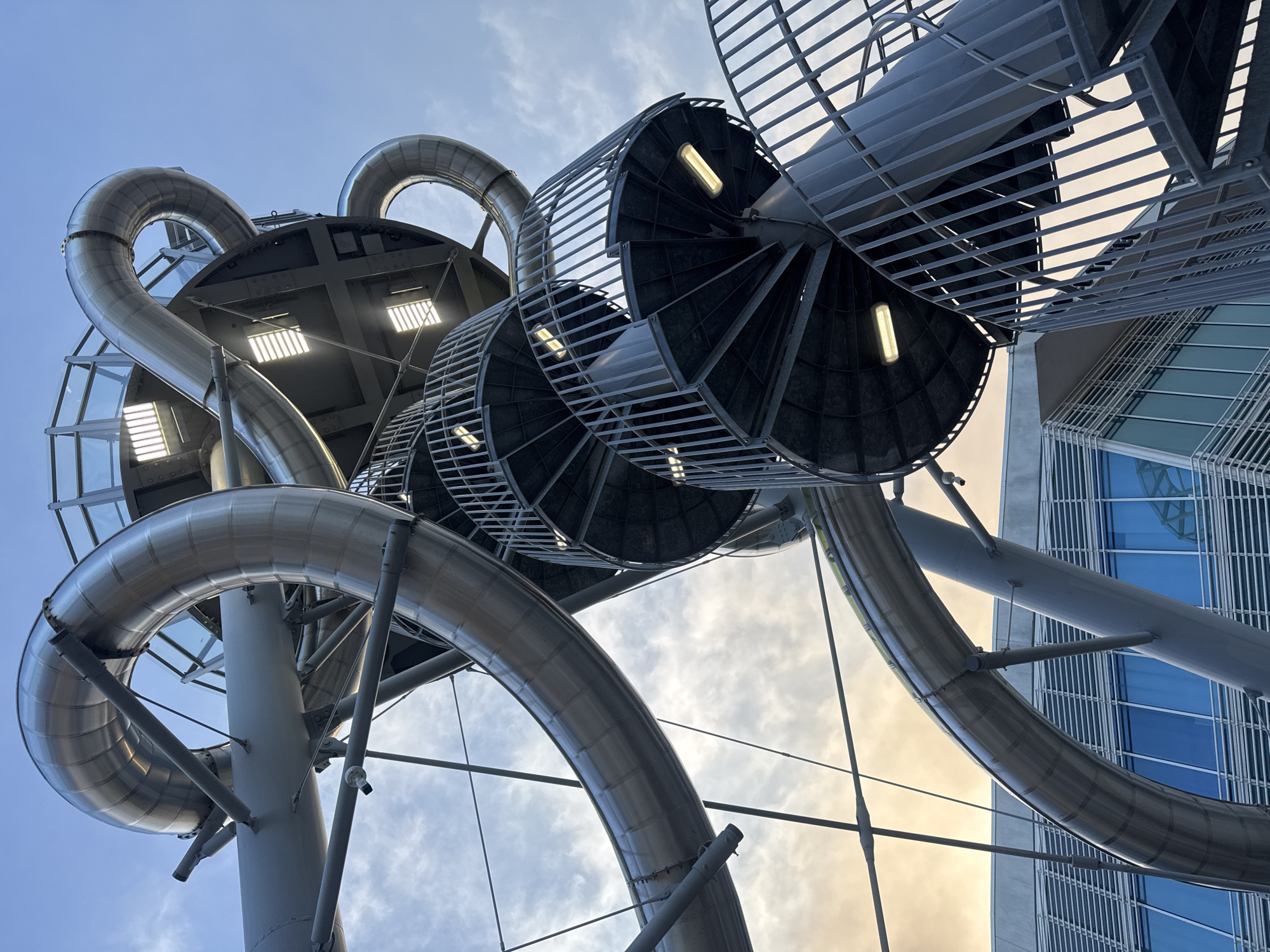 An eye-level, low-angle shot captures a portion of a large, metallic, spiral slide structure against a cloudy sky at dusk. The structure is predominantly silver and gray, with curved, tubular sections of the slide winding upwards and around. Several spiral staircases with metal railings are visible, ascending the structure. The bottom right portion of the image shows a modern building with blue windows and horizontal silver lines. The overall impression is one of industrial architecture and a recreational attraction.