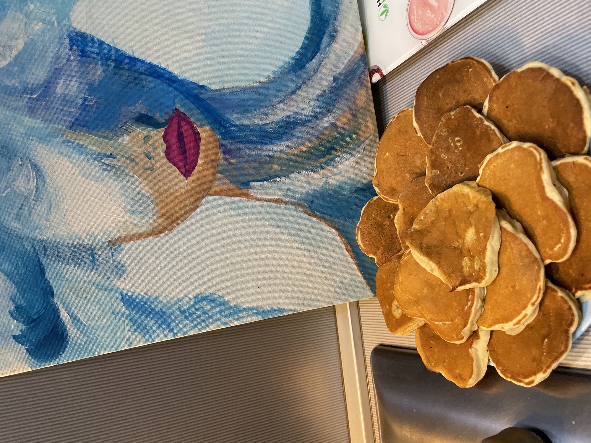 In Sochi, Russia, the scene captures a still life composed of a painting and food arranged on a surface. A vertical canvas painting, depicting a stylized female face, dominates the upper half of the frame, leaning against a light grey, vertically ribbed surface. The painting features vibrant blue and white brushstrokes that suggest hair or an ethereal aura, partially obscuring the face. Prominent magenta-colored lips are visible on the depicted face, which has a warm, light brown skin tone. To the left of the painting, a dark grey, vertically fluted panel forms the background. In the lower half of the image, a white plate contains a pile of approximately 15-20 small, round pancakes. These pancakes exhibit a golden-brown cooked surface with lighter, slightly raised edges. Adjacent to the pancakes on the immediate left, a dark, reflective surface, possibly part of a kitchen appliance, is visible. To the right of the pancakes and partially beneath the painting, a white rectangular packet or container with a green "Herbalife" logo and a circular pink graphic is present on the striped surface.