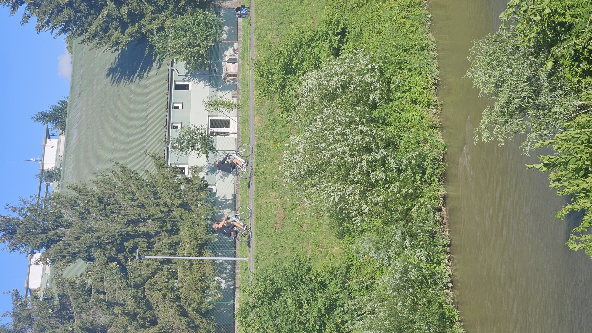 The image captures a sunny day scene in Prešov, Slovakia, featuring a river in the foreground and a building with a green roof in the background. Two individuals are seen cycling on a paved path alongside a grassy embankment. The building, partially obscured by evergreen trees, has a light green facade with multiple windows. A dog kennel structure is visible near the building. The river flows gently, with lush green vegetation lining its banks and extending up the embankment. The sky is clear blue.