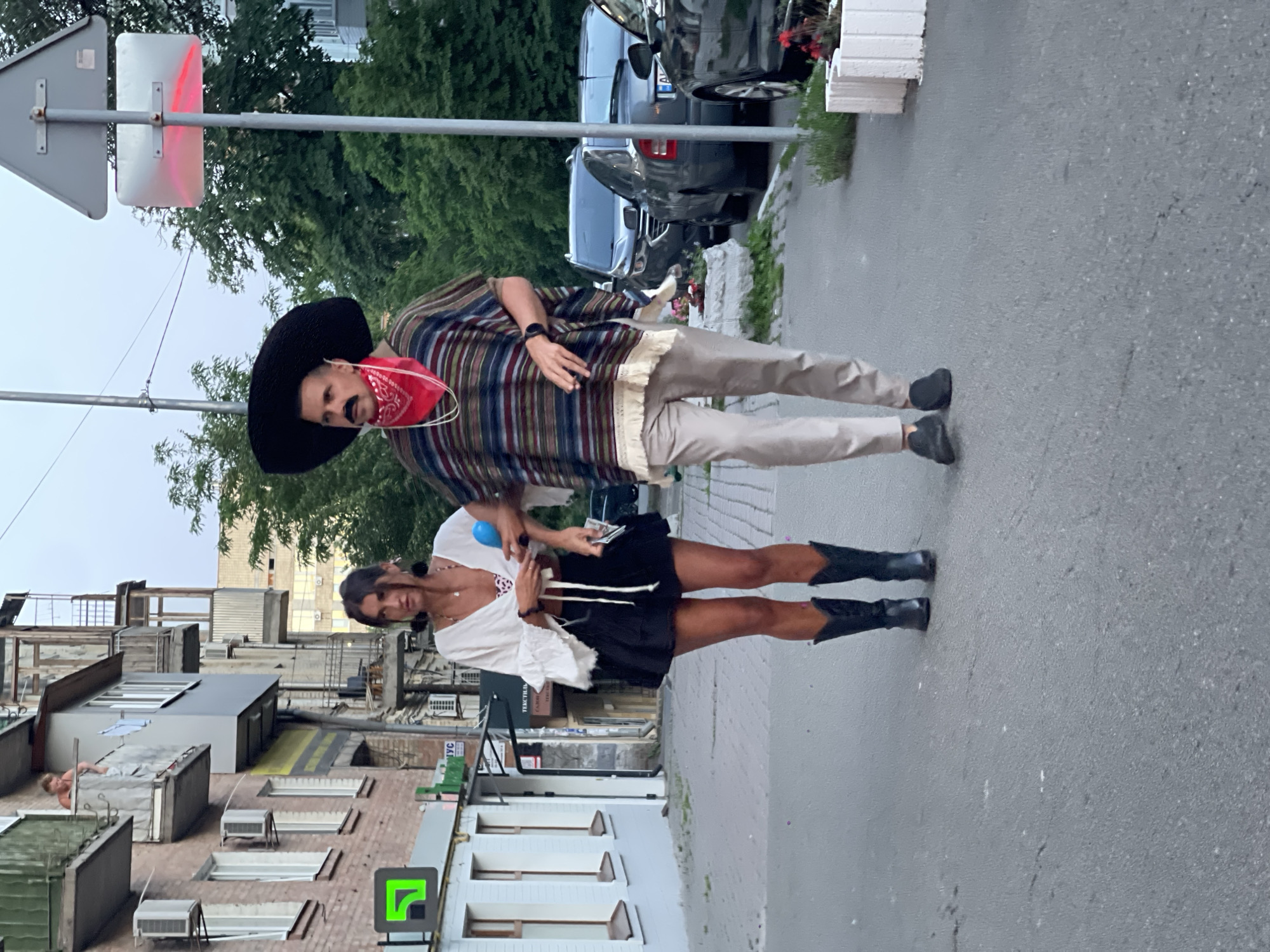 A man and a woman are walking down a sidewalk in Kyiv, Ukraine. The man is dressed in a sombrero, a red bandana, a striped poncho, and tan pants. He has a fake mustache. The woman is wearing a white top with ruffled sleeves, a black skirt, and black cowboy boots. They are walking away from the camera, towards a street with parked cars. In the background, there is a brick building with balconies and trees. The sky is overcast.