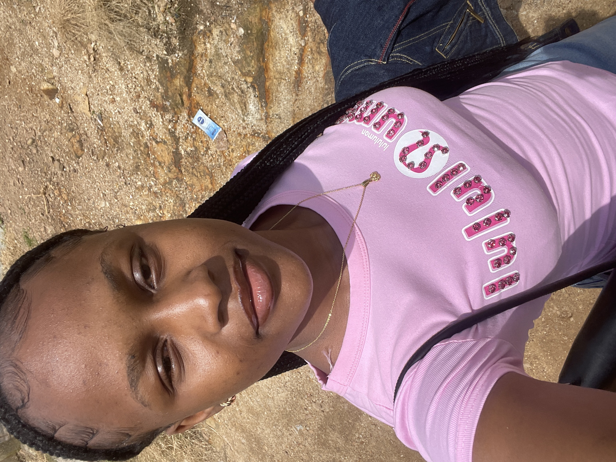 In the city of Rafin Par, Nigeria, a young Black woman with braided hair is captured in a close-up, high-angle selfie outdoors. She is wearing a pink t-shirt adorned with sparkling pink lettering that spells out "Lululemon." A delicate gold necklace with a small pendant rests on her collarbone. The background consists of a textured, dry, rocky ground with sparse vegetation and a discarded blue and white packet. Her expression is neutral, and the lighting suggests it is a sunny day. To her right, a portion of a dark denim jacket is visible.