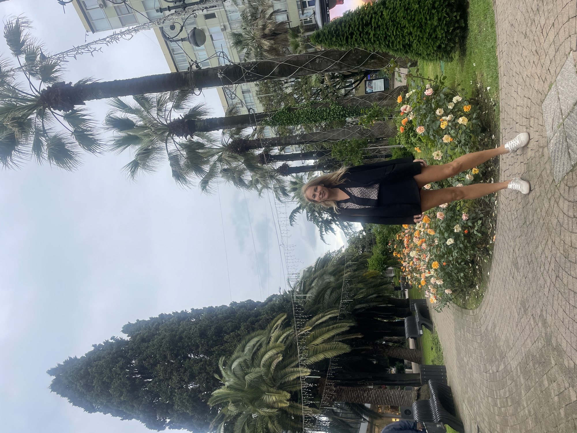 A woman in a black blazer stands in a park setting in Sochi, Russia. She is smiling at the camera. The background includes palm trees, flower bushes with orange and white flowers, a brick pathway, benches, and buildings. The sky is overcast.