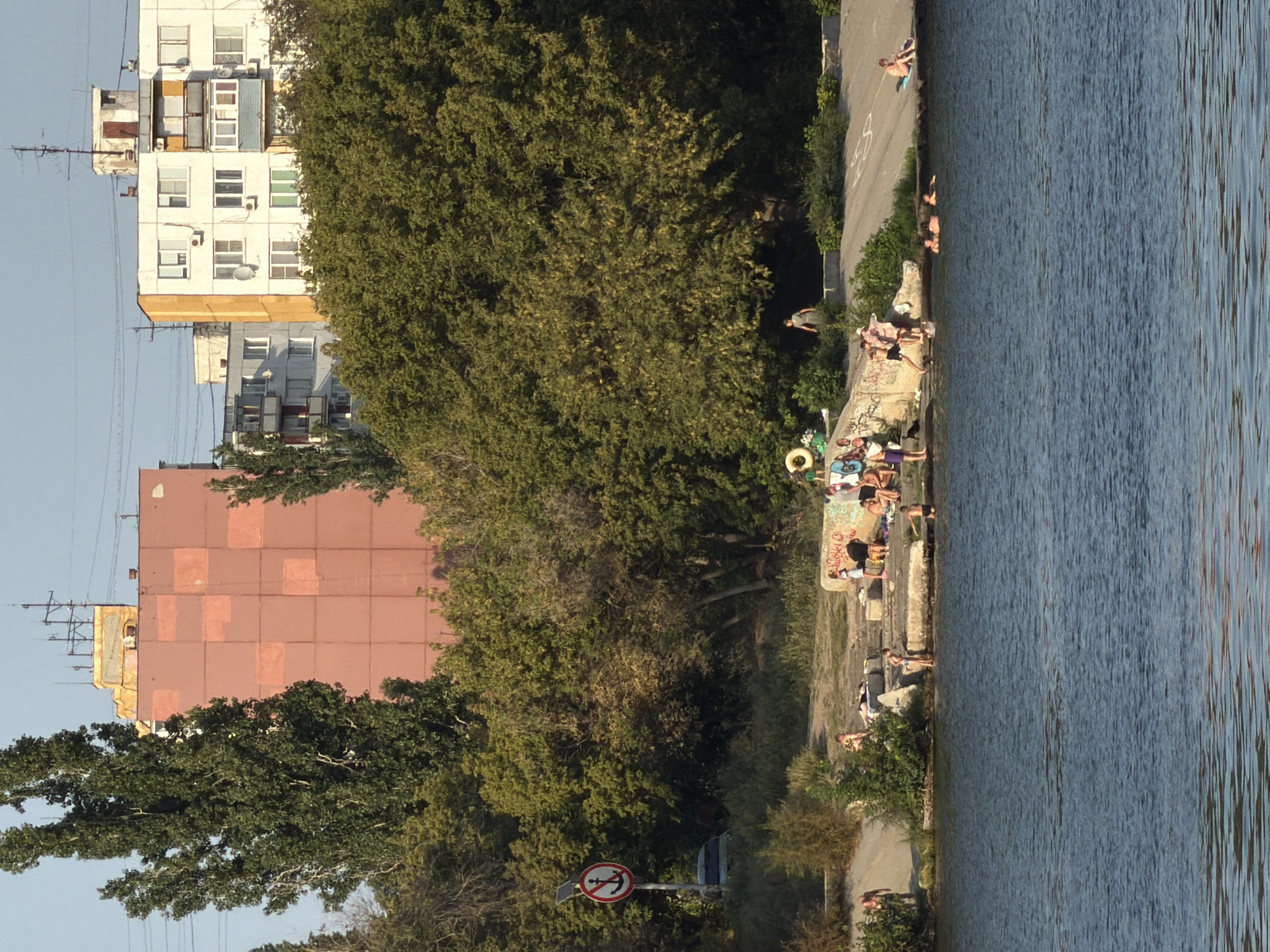 The image captures a sunny day at a body of water, likely a river or lake, in Kamyshin, Russia. In the foreground, the rippling blue water occupies a significant portion of the frame. Along the bank, a concrete embankment is visible, adorned with graffiti. A group of people are gathered on and around this embankment, some sunbathing on towels, others in conversation, and a few are partially submerged in the water, swimming or wading.
Further back, lush green trees dominate the middle ground, obscuring much of the urban landscape behind them. Peeking through the foliage are the upper floors of several apartment buildings. One prominent building on the left is a tall, salmon-pink structure with a grid-like pattern of lighter squares. To its right, a white apartment building with yellow accents stands, featuring multiple balconies and air conditioning units. The sky above is a clear, pale blue, suggesting a warm, cloudless day.
On the left side of the embankment, a sign with a red circle and a diagonal line through an anchor symbol is posted on a pole, indicating a prohibition, possibly related to boats or anchoring. A partially visible car is parked behind some bushes in the distance. The overall scene depicts a relaxed, recreational atmosphere with people enjoying a summer day by the water in a residential area of the city.