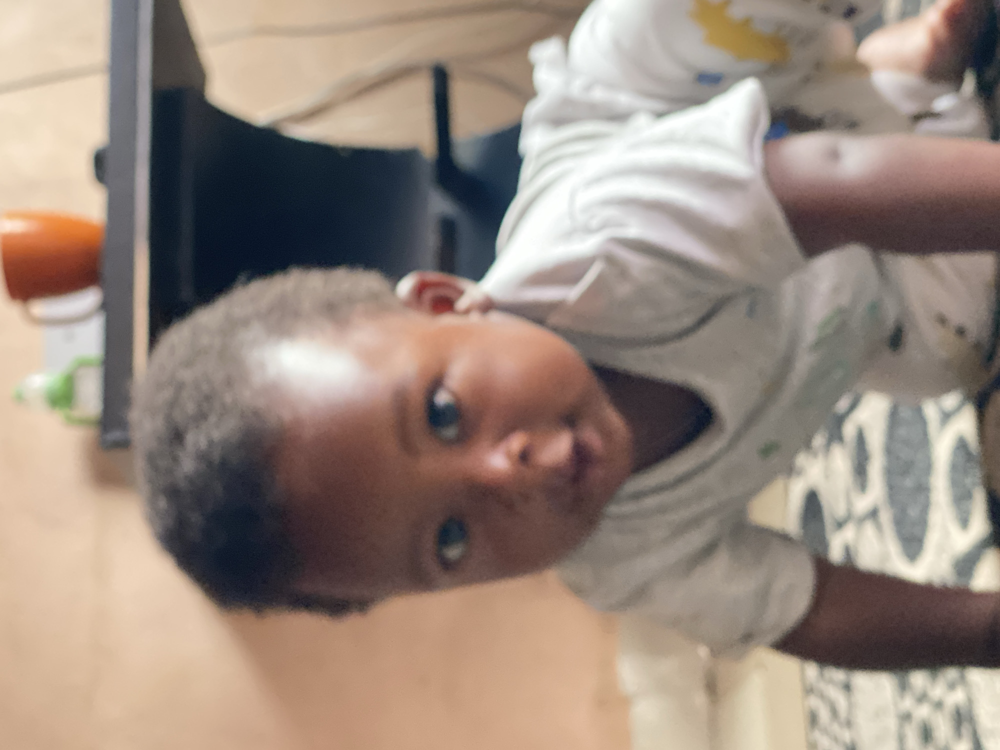 A child is crawling on a patterned rug in an indoor setting. The child is wearing a white outfit with colorful accents. The background appears to be a room with a shelf holding a coffee mug and other items. The overall scene suggests a domestic environment in Sabon Tasha, Nigeria.