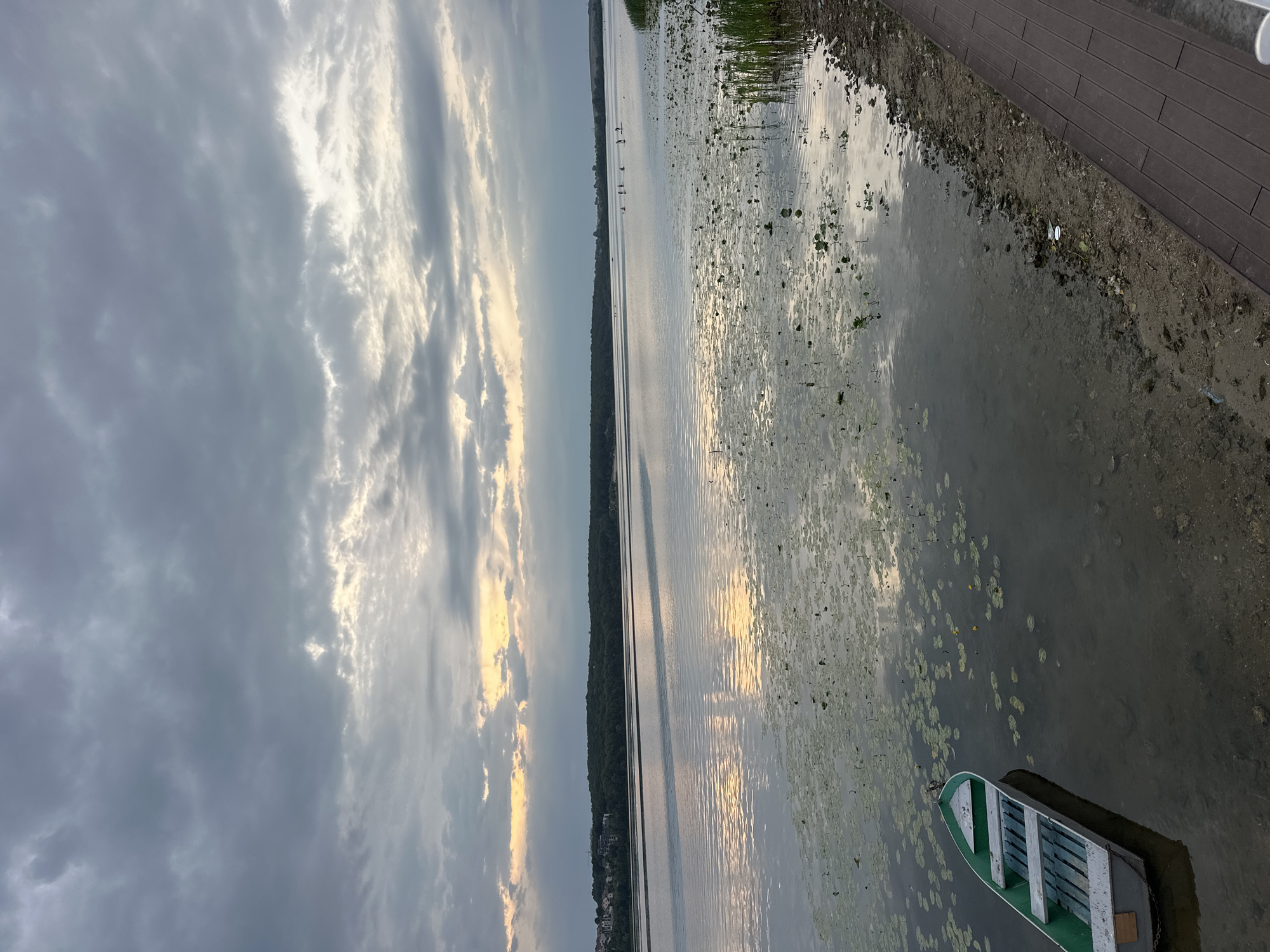 The image depicts a serene lake scene under a cloudy sky. In the foreground, a small green and white rowboat is moored near the shore, surrounded by lily pads. The lake stretches towards the horizon, where a line of trees borders a distant town. The water's surface reflects the subdued light filtering through the clouds. A wooden boardwalk runs along the right edge of the image, indicating a developed recreational area. The overall atmosphere is peaceful and tranquil, characteristic of a lakeside setting in Ternopil, Ukraine.
