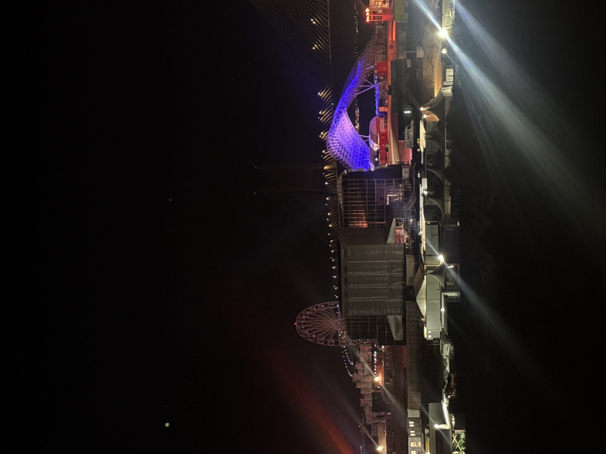 The image depicts a nighttime scene at an outdoor event or construction site in Loures, Portugal. In the background, a large Ferris wheel is illuminated and appears to be part of a temporary setup. A suspension bridge is visible, with its cables and lighting system stretching across the frame. A striking blue, illuminated arched structure stands out on the right side of the bridge. In the foreground, several temporary structures, possibly tents or modular buildings, are set up with artificial lighting. Beams of light from various sources cut through the darkness. A white van with the logo "GMTS" is parked near the tents. The overall impression is of a large-scale operation, possibly an event venue or infrastructure project, at night.