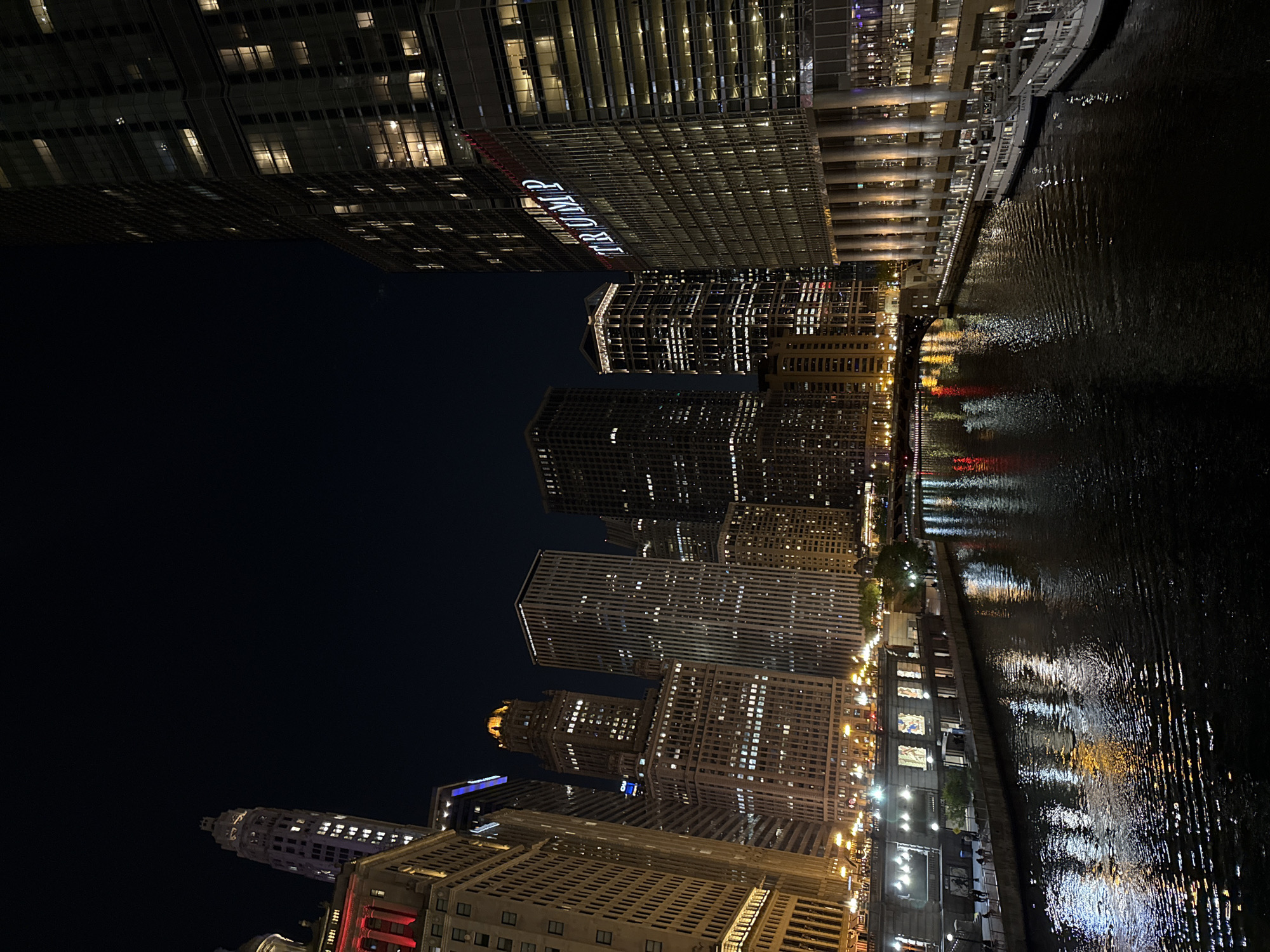 The image shows a nighttime cityscape of Chicago, Illinois, United States. The most prominent feature is the river reflecting the lights of the surrounding buildings. The Trump International Hotel and Tower is visible, along with several other skyscrapers. The buildings are lit up, creating a dynamic play of light and shadow on the water. There are no people visible in the image. The scene depicts an urban environment at night, with a focus on the architecture and the riverfront.