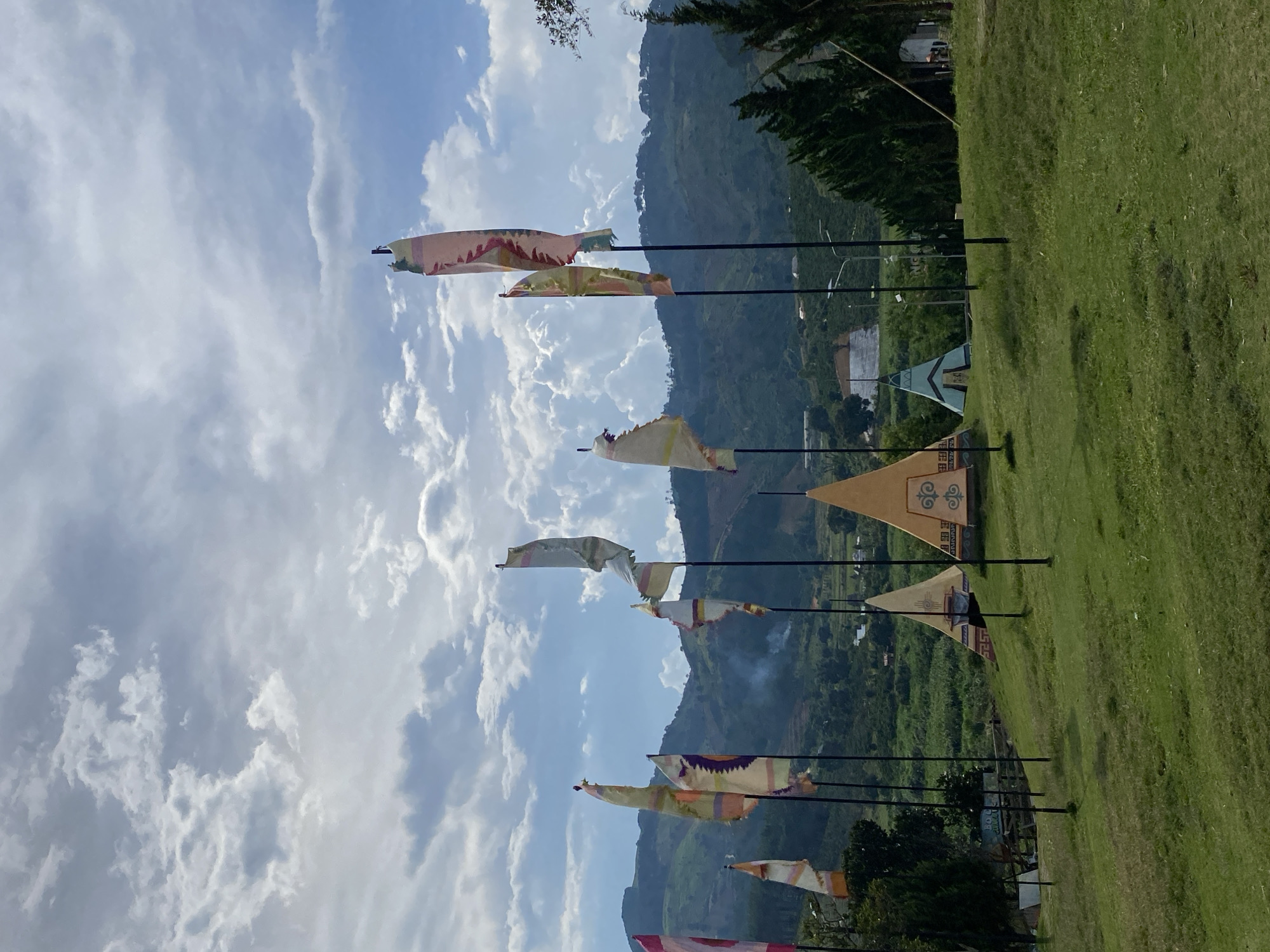 This image depicts a scenic outdoor landscape with rolling hills and a cloudy sky. In the foreground, a grassy hill is dotted with several tall, thin flags on poles, each adorned with colorful patterns. Behind the flags, several teepee-like structures are visible, varying in size and color. The backdrop consists of lush green hills, some of which are terraced, and a few scattered buildings. The overall impression is of a recreational or tourist area situated in a natural, mountainous setting, likely located in or near Thôn 5, Vietnam, given the hilly terrain and vegetation common to the region. There are no people visible in the image.