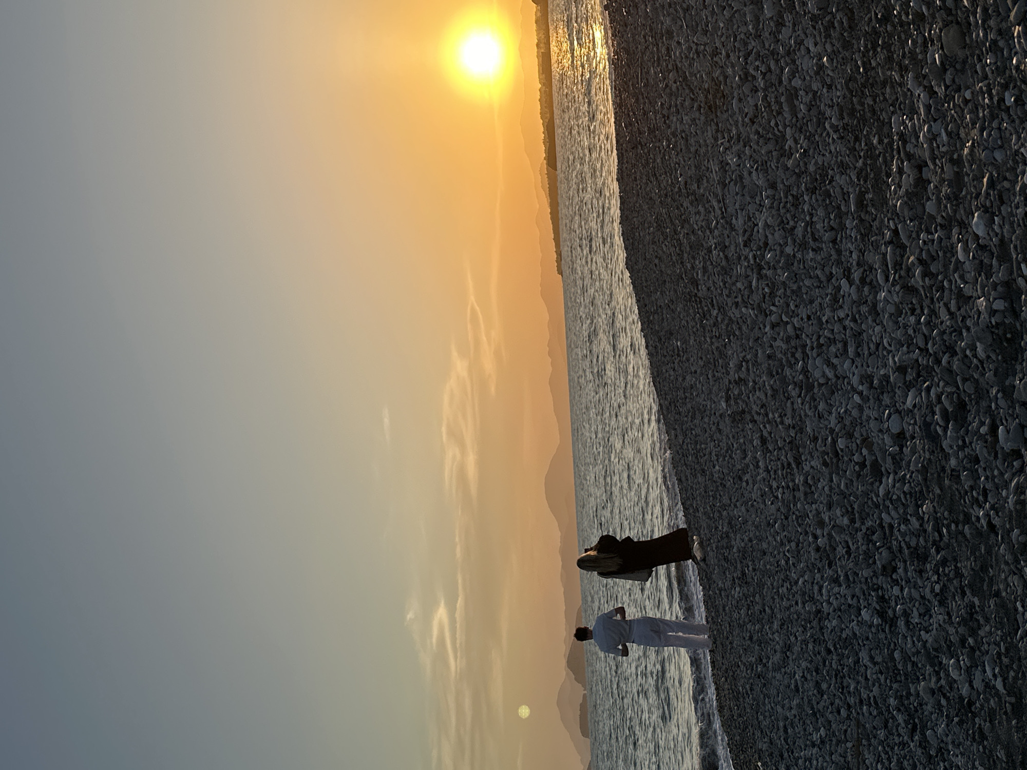 The image shows a beach scene at sunset in Fethiye, Turkey. A man in white clothing is standing on the shoreline. Nearby, a dark-colored object is lying on the beach, possibly a jet ski or another watercraft. The beach is covered in pebbles. The sun is setting over the mountains in the distance, casting a warm golden light on the sky and sea. The sea appears calm, with gentle waves lapping at the shore.