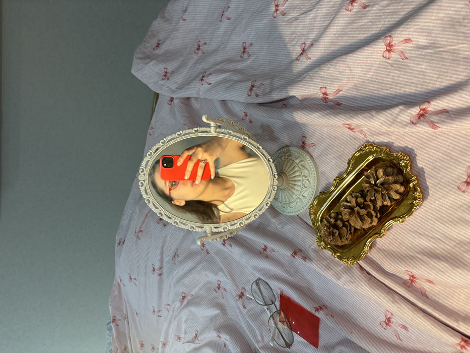 The image displays an indoor scene, likely a bedroom, where a bed is covered with light pink and white striped bedding patterned with red bows. A white, ornate, oval-shaped vanity mirror with a stand is centrally positioned on the bed. Reflected in the mirror is the upper body of an individual. This individual holds a bright red smartphone with their right hand, covering the lower portion of their face, and wears red-framed eyeglasses and a light-colored top. A ring is visible on the ring finger of the hand holding the phone. To the left of the mirror, a pair of eyeglasses with thin metal frames rests on a small, rectangular red object. In the lower right quadrant of the image, a small, gold-colored, ornate rectangular tray contains three pinecones. A light blue-grey wall is visible in the background above the bed. The specific location within the city of Krasnodar, Russia, is not discernible from the provided visual information.