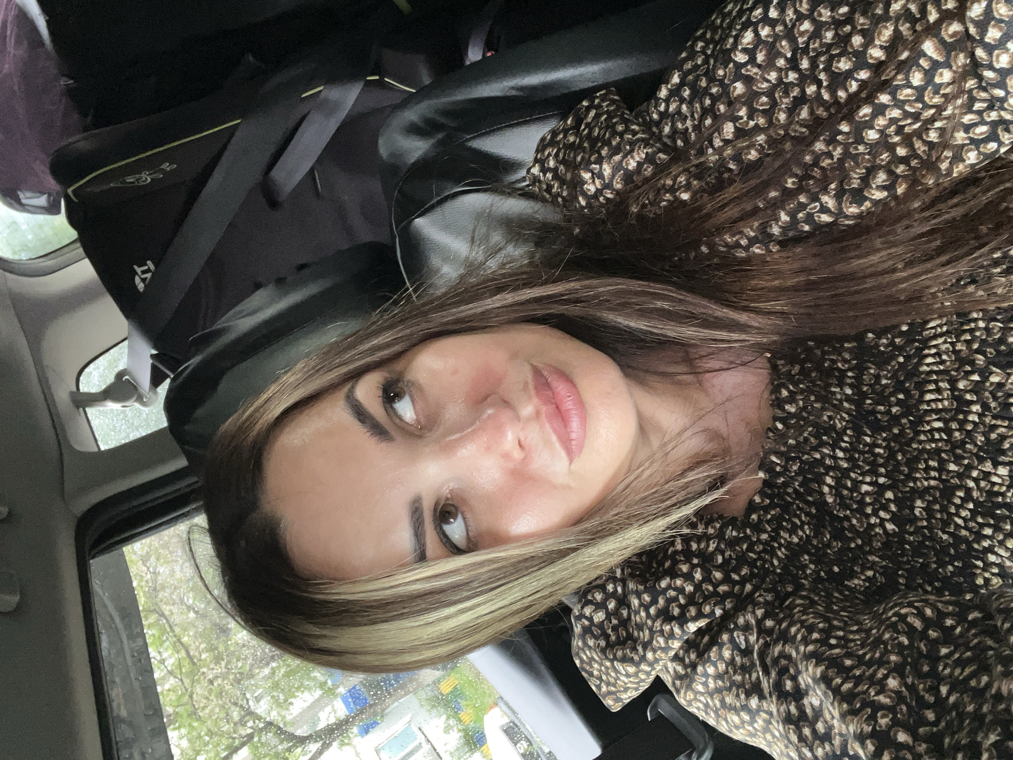 A woman with long, dark brown and blonde streaked hair is seated in the passenger seat of a car, looking to her right and slightly upwards. She is wearing a brown and black leopard-print top. The background shows a car seat, a car window with trees and buildings visible outside, and other car interior elements. The weather appears to be overcast and possibly raining, as evidenced by water droplets on the car window.