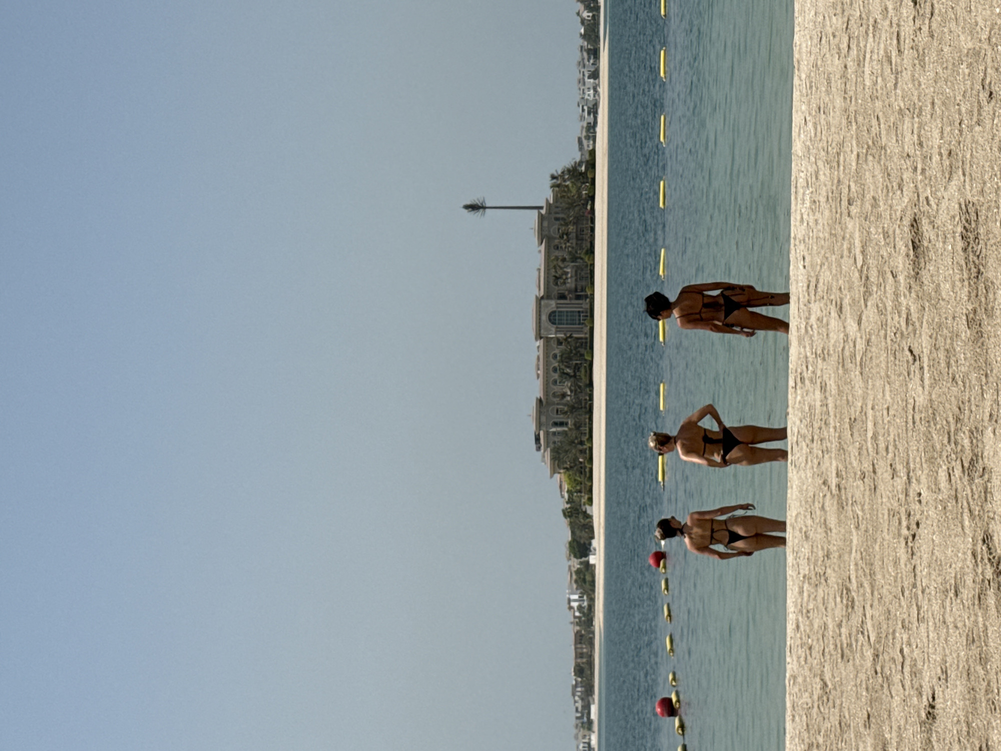 Three women, viewed from behind, stand in shallow ocean water at a beach in Dubai, United Arab Emirates. They are all wearing bikinis. The woman on the left has dark hair, the woman in the middle has blonde hair, and the woman on the right has dark hair styled in a bun. In the background, a large, ornate building with a tall, slender tower in front of it is visible, situated on a sandy coastline with palm trees. Yellow buoys and red spheres float on the water, marking a boundary. The sky is clear and blue.