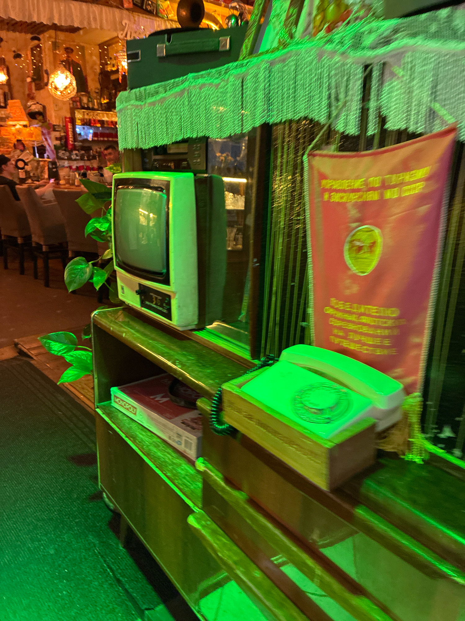 The image depicts a dimly lit interior space, possibly a bar or restaurant, illuminated by vibrant green lighting and a warm, golden glow from decorative lamps. In the foreground, a vintage green television set sits atop a wooden shelf unit. To the right of the television, a white rotary-style telephone rests on a wooden surface. Behind these objects, a red banner with Cyrillic text is partially visible. A Monopoly board game box is also present on the shelf unit. In the background, seating areas with chairs and tables are visible, along with shelves stocked with bottles, suggesting a bar setting. Several people are subtly present in the background, some seated at tables or the bar. The overall ambiance suggests a retro or themed establishment. The specific location in Sochi, Russia, cannot be determined from the visual information provided.