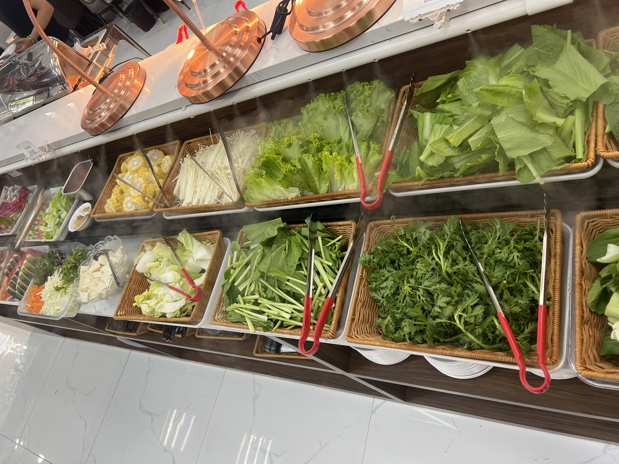 A high-angle shot depicts a salad bar, likely in a restaurant, featuring a variety of fresh vegetables and some other food items arranged in woven baskets and trays. The lighting is bright, with some steam or mist suggesting temperature control or freshness.
From left to right, one can see trays containing items like sliced dragon fruit, lettuce, carrots, tofu cubes, cabbage, corn on the cob, enoki mushrooms, various leafy greens, and other vegetables. Tongs with red handles are placed in each basket or tray, presumably for customers to serve themselves.
Above the food, there are dome-shaped copper-colored fixtures, possibly heat lamps or decorative elements. The salad bar is positioned against a white tiled floor and a clean countertop.
While the image doesn't provide explicit clues about the city, given the style of cuisine often found in Vietnam and the prevalence of fresh vegetables, it's plausible that this salad bar could be located in Đà Nẵng.