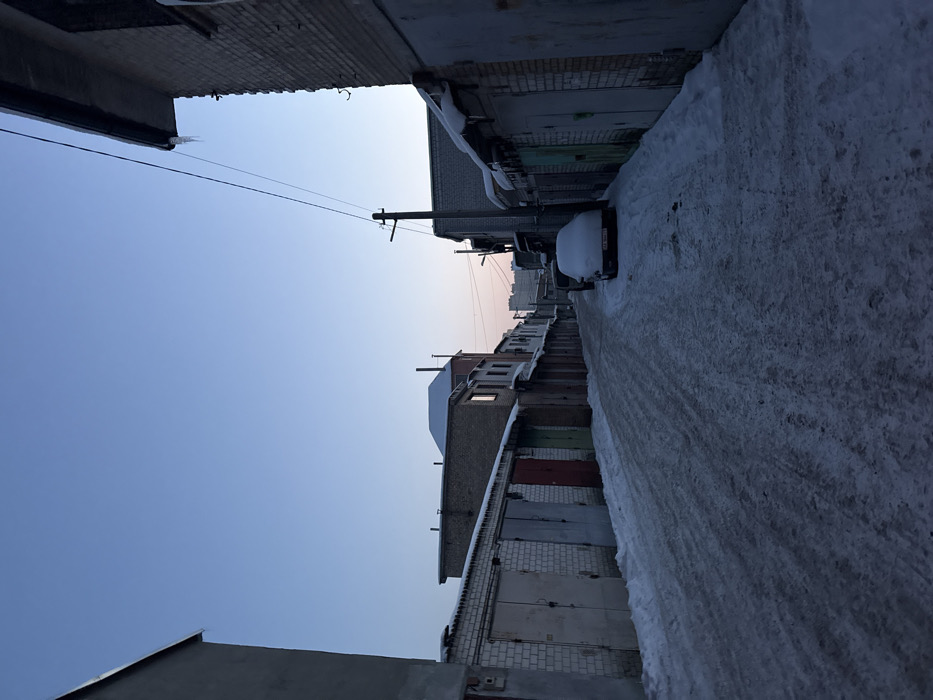 A winter alley scene with snow-covered ground. Buildings line the sides, featuring garage doors. A car, also covered in snow, is parked along the alley. Power lines stretch across the clear sky above. The picture is taken at an angle, emphasizing the perspective along the alley. The sky is blue with a hint of sunrise/sunset light.