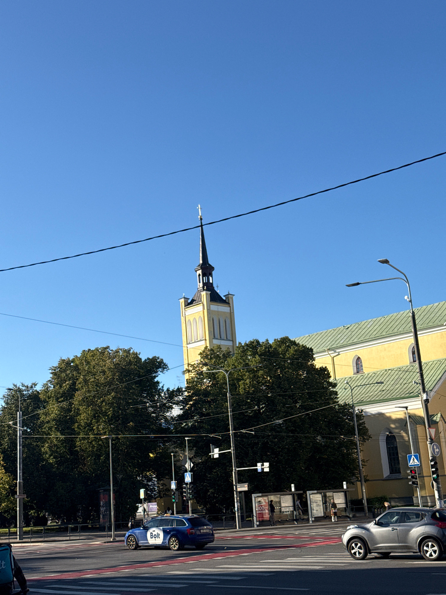 The image shows a view of St. Olaf's Church (Oleviste kirik) in Tallinn, Estonia, on a clear day with a blue sky. A portion of the church's yellow facade and green roof is visible on the right side of the frame, along with its tall, dark spire topped with a cross. Dense green trees partially obscure the lower parts of the building.
In the foreground, a busy intersection is depicted with pedestrian crossings marked by red and white stripes. A blue Bolt car, a ride-sharing service, is driving through the intersection. To the right, a silver car is also navigating the intersection. Several streetlights and traffic signals are present. People are seen waiting at bus stops and walking on the sidewalks. Overhead power lines are visible across the upper part of the image.