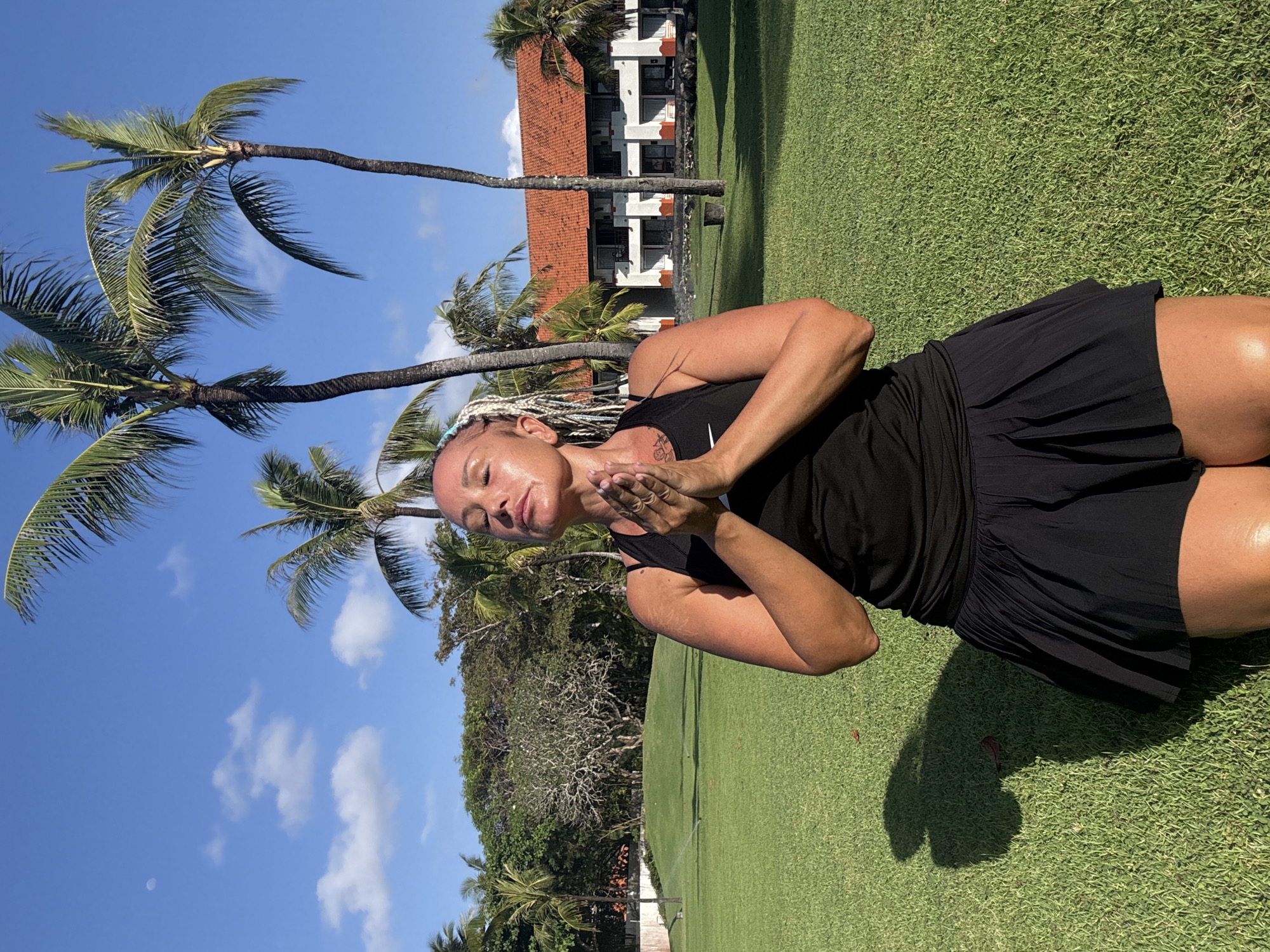 A woman with light-colored braided hair is captured in a vertical shot, standing on a vibrant green lawn under bright sunlight. She is wearing a black tank top and a black pleated skirt. Her eyes are closed, and her hands are pressed together in front of her chest in a prayer or meditative gesture (Anjali Mudra). A small, white logo, possibly a Nike swoosh, is faintly visible on her tank top near her left collarbone. On her left upper arm, there is a tattoo with an abstract design or script. Strong shadows from the woman are cast on the grass to her right, indicating a sunny day, likely midday or early afternoon.
The background features several tall palm trees with green fronds, characteristic of a tropical climate. Behind the trees, a multi-story building with a distinctive red-tiled roof and white facade is visible. The sky above is clear blue with a few scattered white clouds, one of which appears to obscure a faint moon. The overall scene suggests a peaceful, contemplative moment in an outdoor, resort-like setting in Kuta Selatan, Indonesia.
