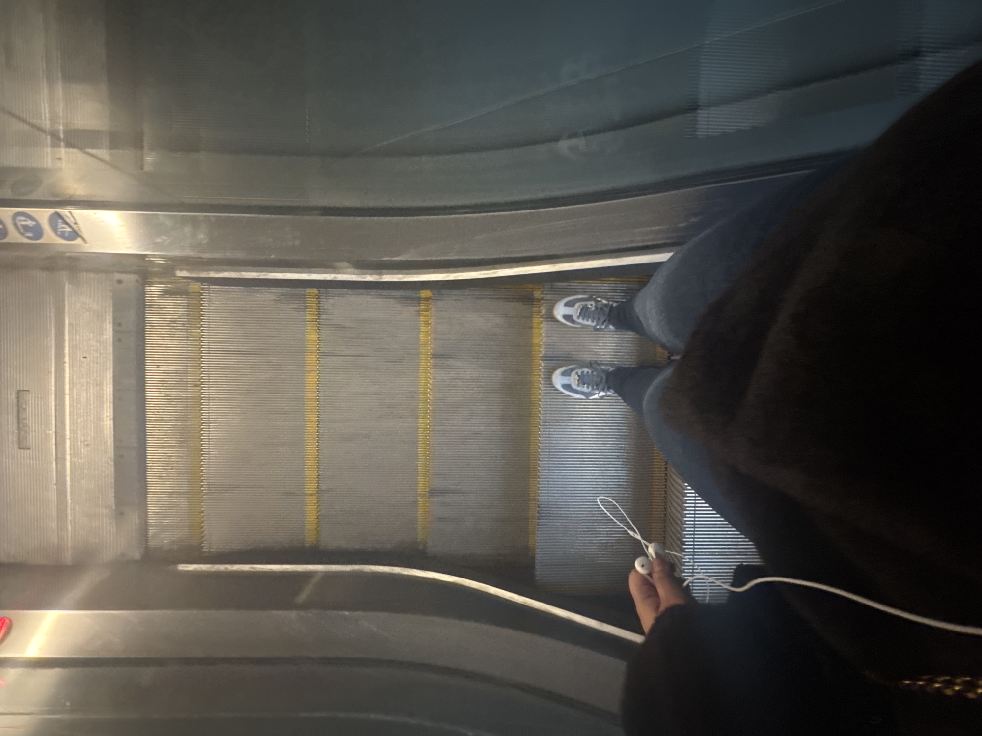 A person stands on an escalator in Sofia, Bulgaria. The person wears jeans and gray shoes with white soles. They hold white earphones. The escalator has a yellow-striped, grooved, silver surface. A black garment partially obscures the person. Signs in Bulgarian are visible on the side of the escalator.