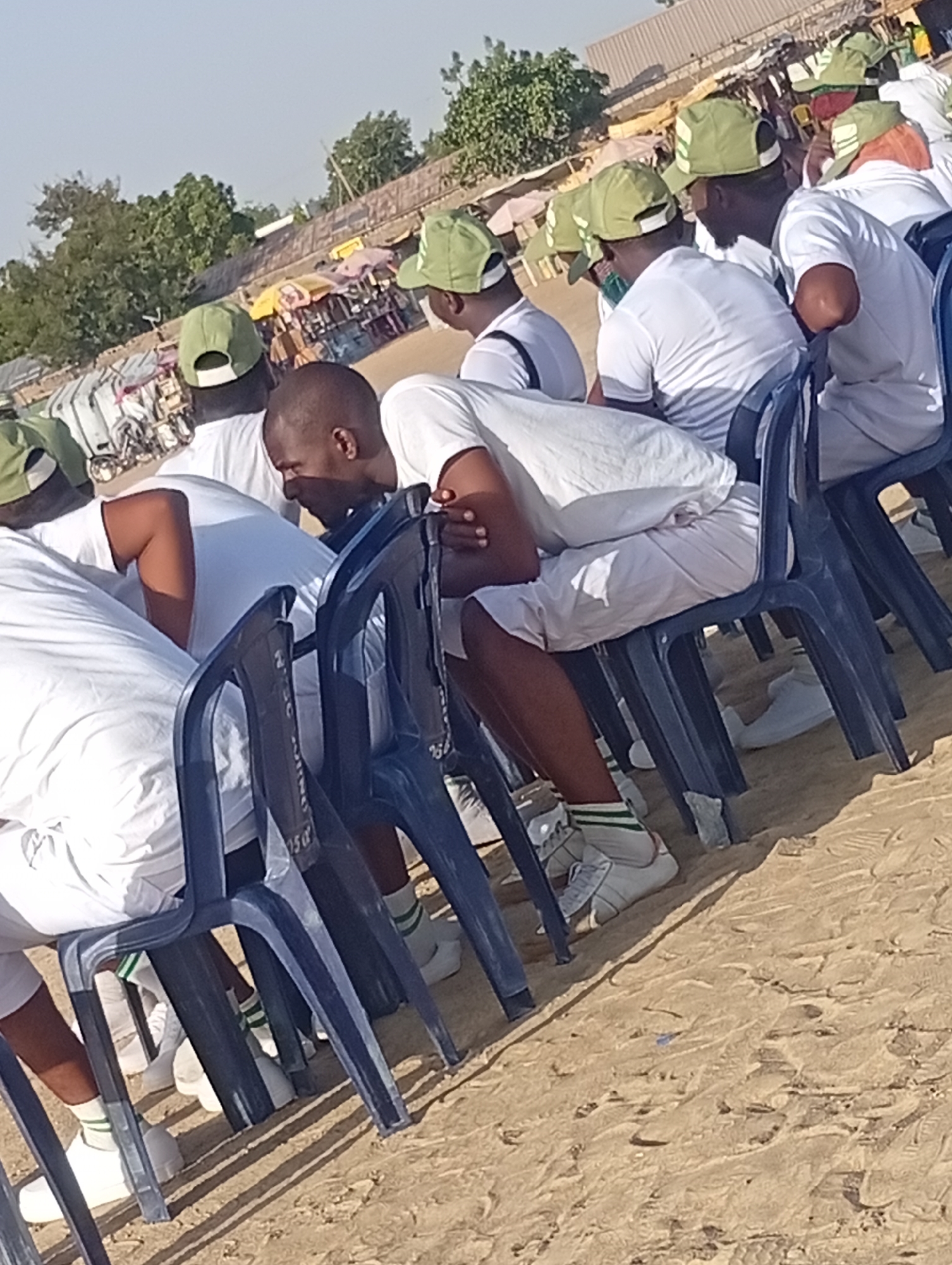 The image shows a group of people, mostly men, seated in blue plastic chairs on a sandy surface. They are uniformly dressed in white t-shirts, white shorts, and white sneakers with green and white striped socks. Most are wearing green and white caps. They appear to be lined up in rows.
In the background, there are hints of a marketplace or street vending area with colorful umbrellas or canopies. There are also trees and buildings visible in the far distance.
The scene seems to be set outdoors in a sunny location. The sandy ground suggests an arid or semi-arid environment. Given the context of the city, the scene is probably set in Maiduguri, Nigeria.