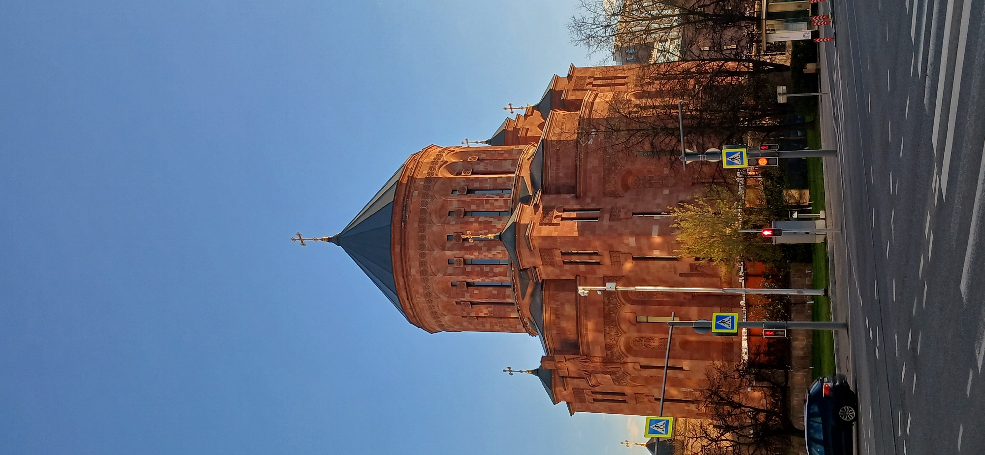 The image shows the Armenian Apostolic Church, Holy Resurrection Cathedral, located in Moscow, Russia. The church is constructed from a reddish-brown stone. A street runs beside the church with pedestrian crossing signs and traffic signals visible. There's a dark-colored car parked on the side of the road. The sky is clear and blue. Trees with bare branches are visible, suggesting it could be late autumn or early spring. In the background, part of a building is also visible, hinting at the urban environment.