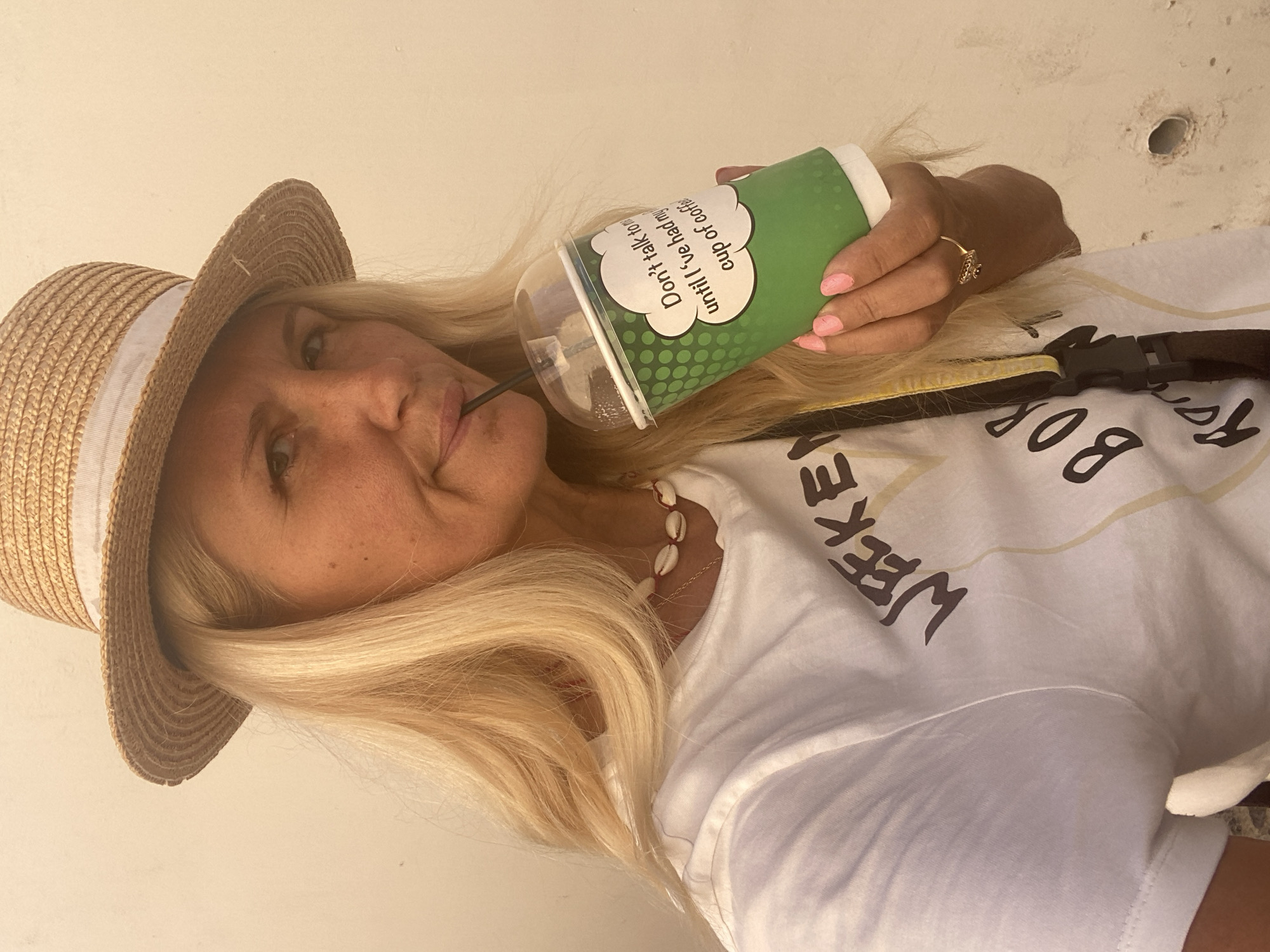 A woman wearing a straw hat and a white t-shirt with a shell necklace is holding a green disposable cup with a straw and drinking from it. The cup has text that reads "Don't talk to me until I've had my cup of coffee". She has blonde hair and is wearing pink nail polish. A ring is visible on her right hand. The background appears to be a light-colored, textured wall, possibly in Ia, Municipal Unit, Greece. The overall lighting suggests an outdoor setting during the daytime.