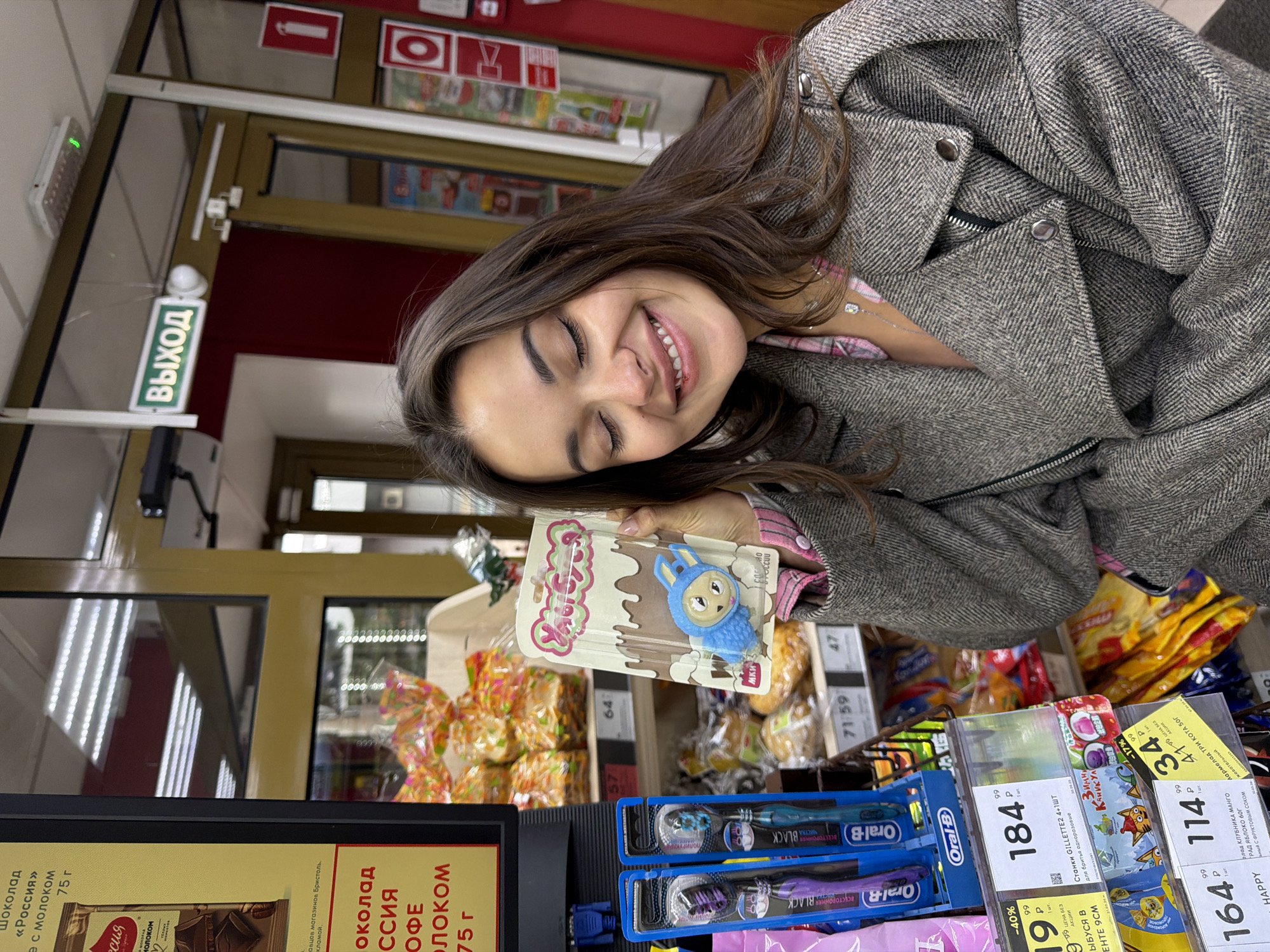 A young woman wearing a grey herringbone coat is smiling and holding a package of "Хрюсka" candy with a cartoon blue bunny on the packaging. She is standing in a store, likely a grocery store or supermarket, looking at the camera. Visible behind her are shelves stocked with candy, toothbrushes (Oral-B Black), and other packaged goods. Pricing labels are visible, with the currency likely in Russian Rubles, as are product descriptions in Russian. An "EXIT" sign is visible in the upper left corner, written in Russian. The location is likely Krasnoyarsk, Russia, based on the presence of Russian text and products commonly found in Russian stores.