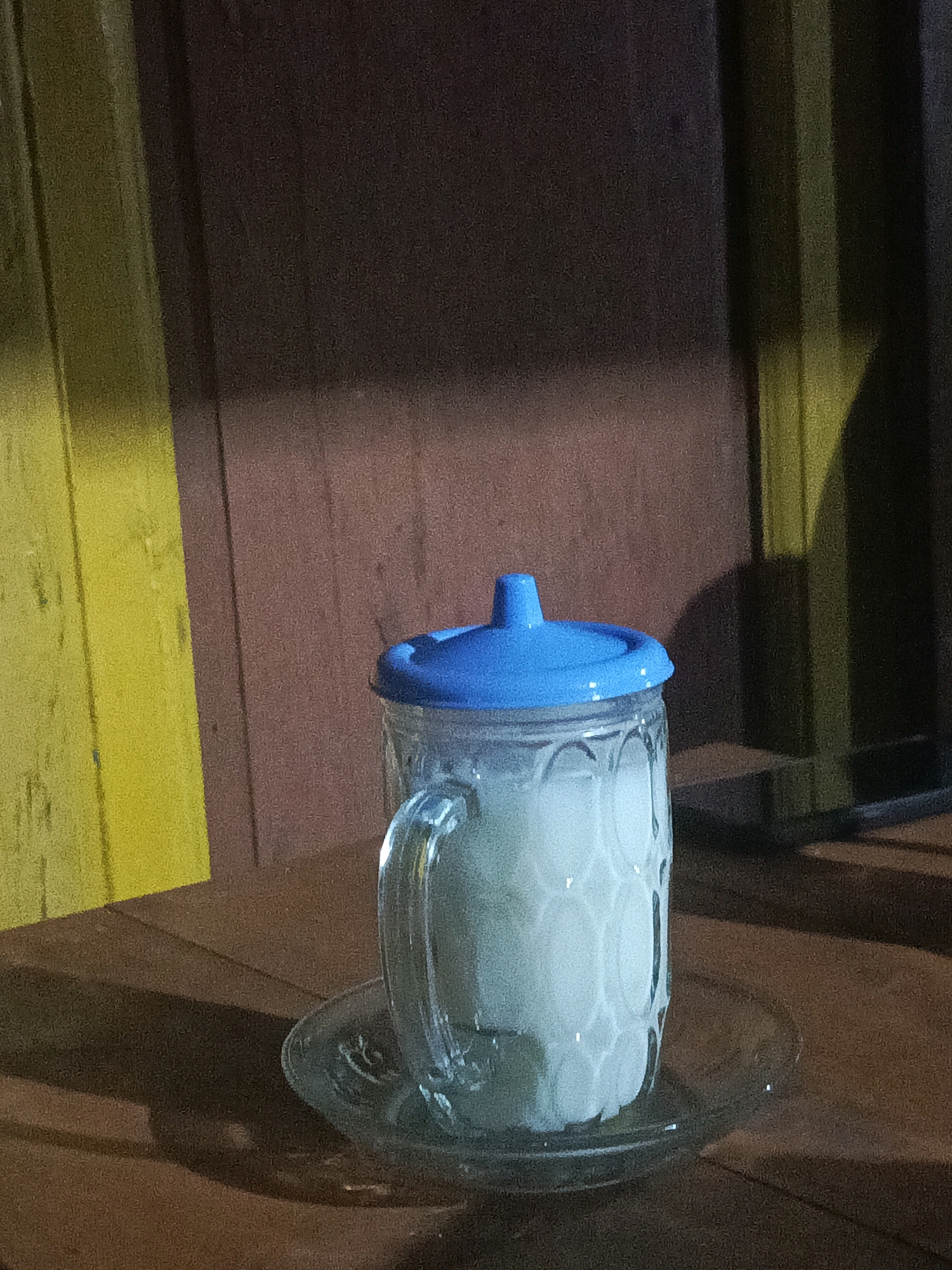 A close-up, low-angle shot captures a glass mug filled with a light-colored liquid, possibly milk or a creamy beverage. The mug has a clear glass body with a textured, patterned surface and a prominent handle. A bright blue plastic lid with a raised spout covers the top of the mug. The mug rests on a clear glass saucer. The setting appears to be outdoors, possibly a warung or food stall in Plumpang, Indonesia, given the wooden wall in the background. The wall exhibits a vertical wood grain texture, with sections painted in yellow and dark brown, and illuminated by artificial lighting that casts distinct shadows. The lighting creates a moody, atmospheric effect, highlighting the mug and its contents. The overall scene is dimly lit, suggesting it might be evening or nighttime.