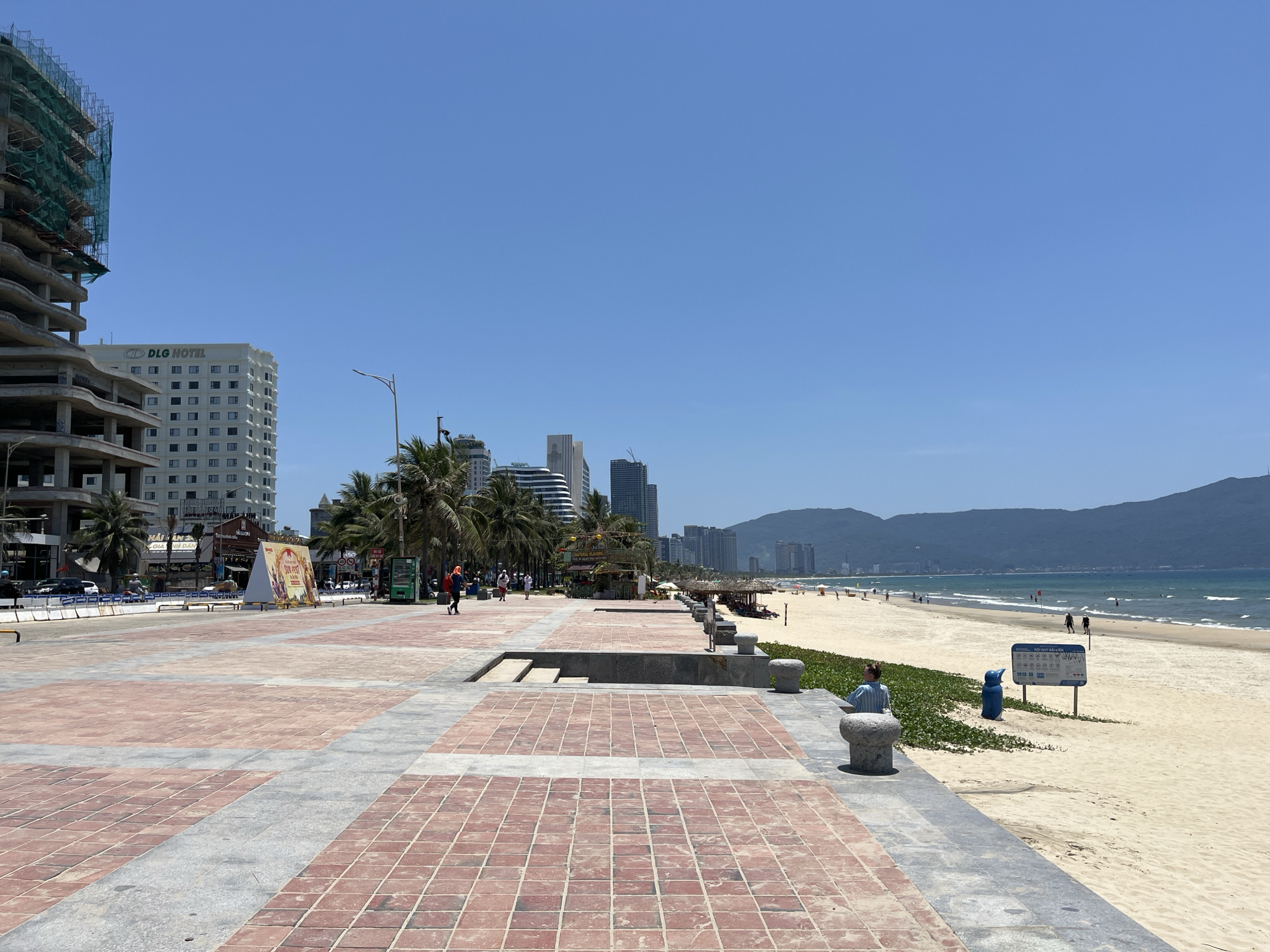 The image depicts a beach promenade in Đà Nẵng, Vietnam, on a clear, sunny day. The foreground is a wide, brick-paved walkway transitioning into a sandy beach. In the midground, there are palm trees, food stalls, and pedestrians. Some people are on the beach near the water. A building with the logo "DLG Hotel" is visible on the left side of the image, along with another building under construction next to it. Mountains are visible in the background across the bay. A blue refuse bin and a white sign with graphic warnings/instructions are positioned on the sand. A person sits on a stone feature that lines the promenade.