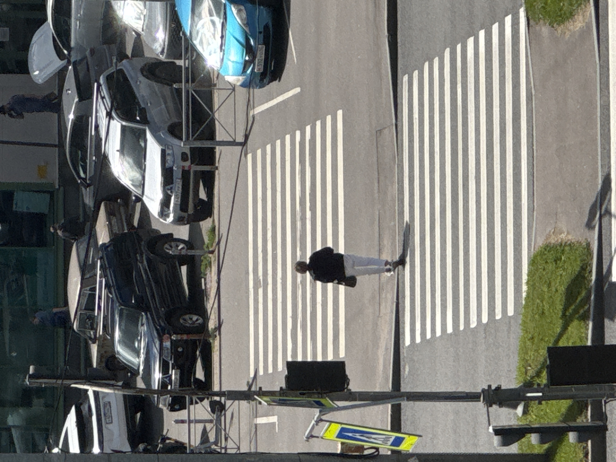 In Saint Petersburg, Russia, on a sunny day, a person walks across a pedestrian crossing on a street. The individual, wearing a dark jacket and light-colored pants, carries a dark bag. They are positioned on the second pedestrian crossing from the top of the frame. Multiple vehicles are parked and moving around the scene. These include a black SUV, a silver SUV, a blue hatchback, and other vehicles partially visible in the background. The environment consists of asphalt roads, sidewalks, grassy areas, and street furniture like lampposts and traffic signs. The buildings in the background are indistinct, suggesting an urban setting.