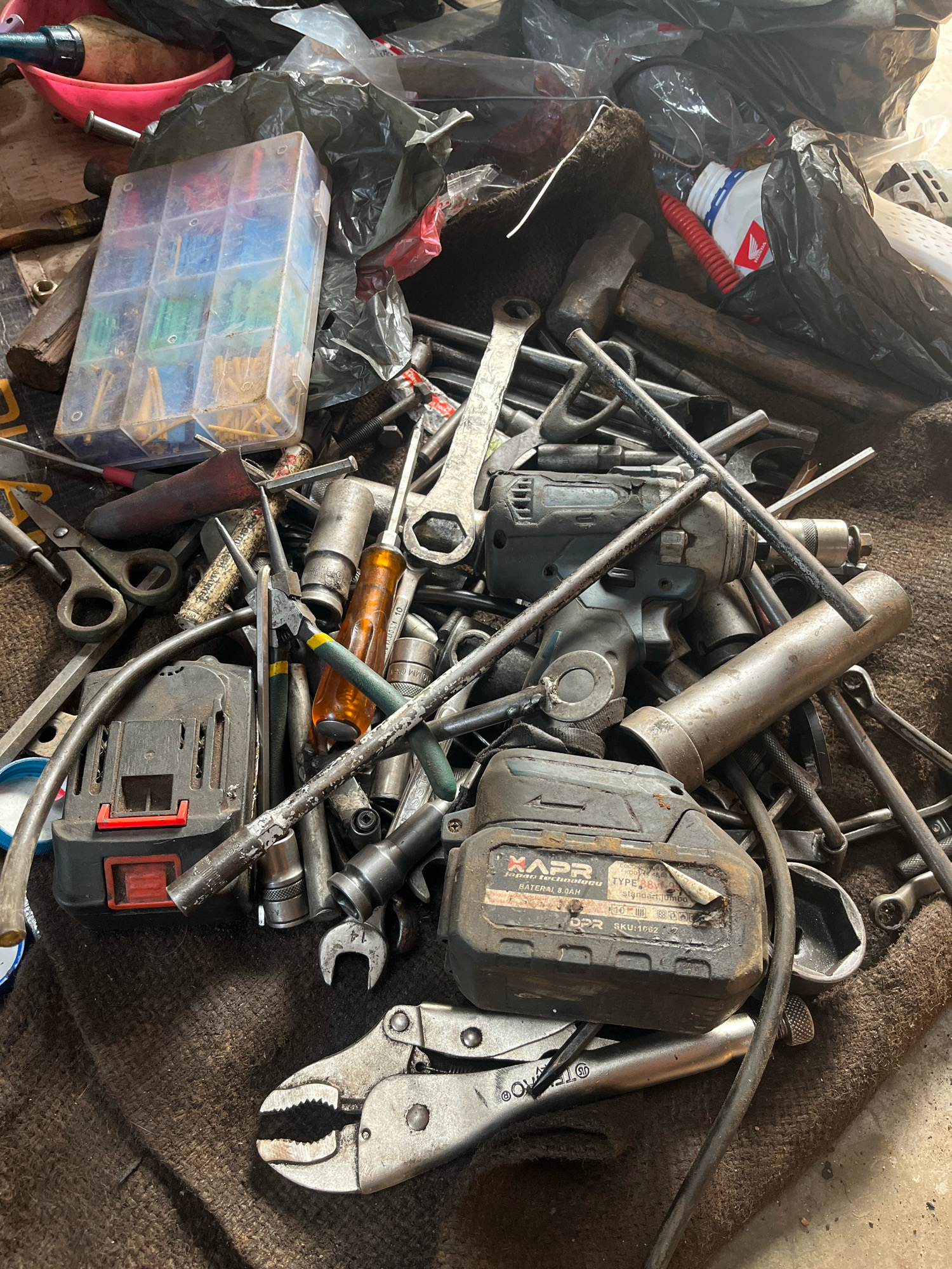 The image depicts a cluttered collection of tools, primarily metal hand tools and a power tool, spread across a brown, textured surface. The tools are a mix of wrenches, sockets, pliers, a hammer, scissors, and a cordless drill with a battery pack. Many of the tools are worn and appear to be in use or storage. A small plastic organizer filled with various colored electrical connectors is also visible. The scene suggests a work area or a storage space for tools, possibly in a garage or workshop. The lighting is overhead and casts shadows, indicating an indoor setting. The specific location in Kebumen, Indonesia, is not identifiable from the image itself, as there are no discernible landmarks or contextual clues.