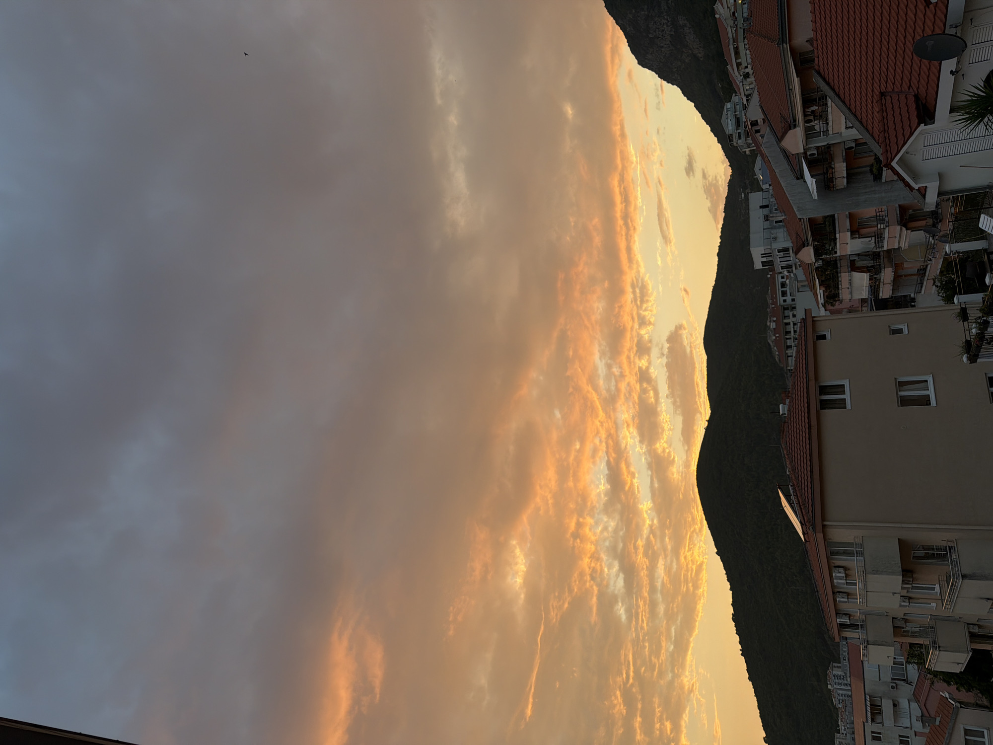 A photograph captures a scenic sunset over the coastal city of Budva, Montenegro. The sky is a dramatic canvas of warm orange and yellow hues, contrasting with darker gray clouds. Below the sky, the silhouettes of rolling hills covered in dense green foliage form a backdrop to the urban landscape. The foreground features a cluster of buildings with terracotta-tiled roofs, typical of Mediterranean architecture. Balconies and windows are visible, suggesting residential or hotel structures. A few scattered buildings are also seen further up the hills, blending into the natural environment. One building in the foreground has solar panels on its roof. The overall impression is one of a tranquil evening settling over a picturesque coastal town.