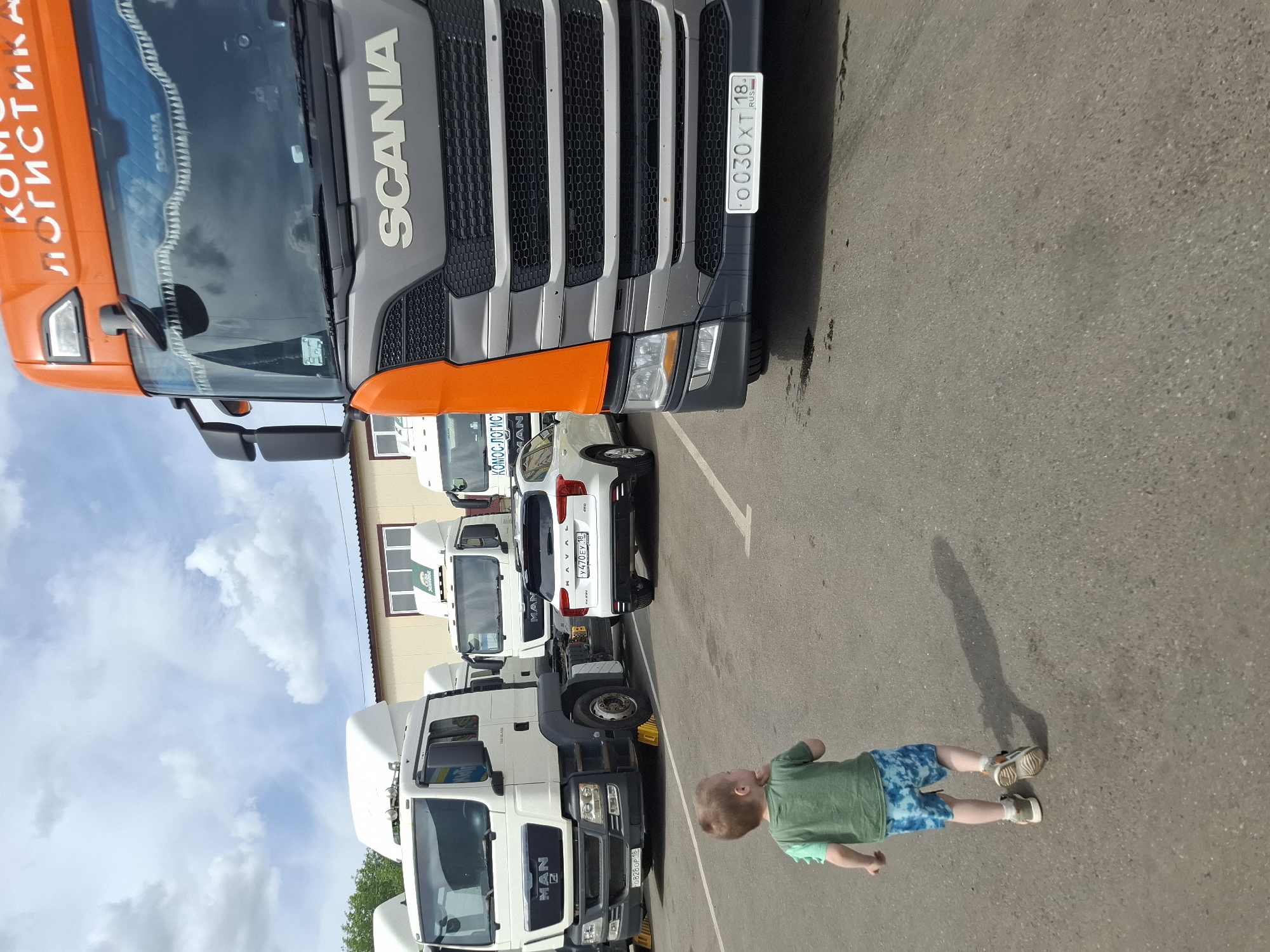 The image shows a parking lot in what appears to be an industrial area. Several large trucks are parked in a row along a building. The trucks include brands like MAN and Scania. A young child is lying on the asphalt surface of the parking lot. The license plates on the trucks and a car indicate the location is in Russia. Specifically, the registration code '18' points to the Republic of Udmurtia, with Izhevsk as the capital city.