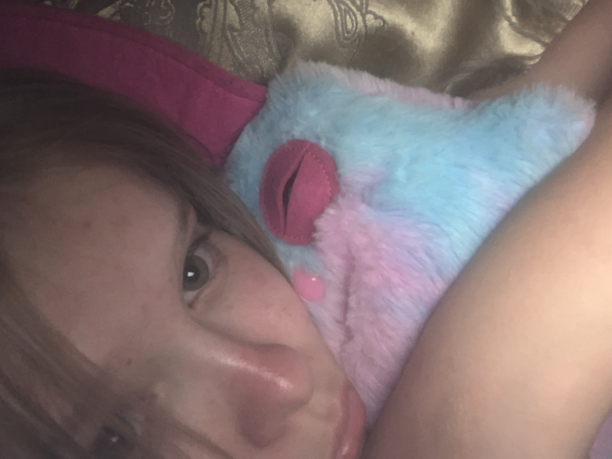 The image is a close-up, low-angle, selfie-style shot of a young person's face and upper body. The person has light-colored hair and fair skin with some redness on their cheeks and nose. Their left eye, which is green, is visible. They are holding a plush toy that is primarily light blue and pink, with a pink mouth detail. The background appears to be a bed with a patterned fabric. The lighting is dim and warm, suggesting an indoor setting. The image does not contain any information that would allow for identification of a specific location within a city, nor does it provide any context related to Russia.
