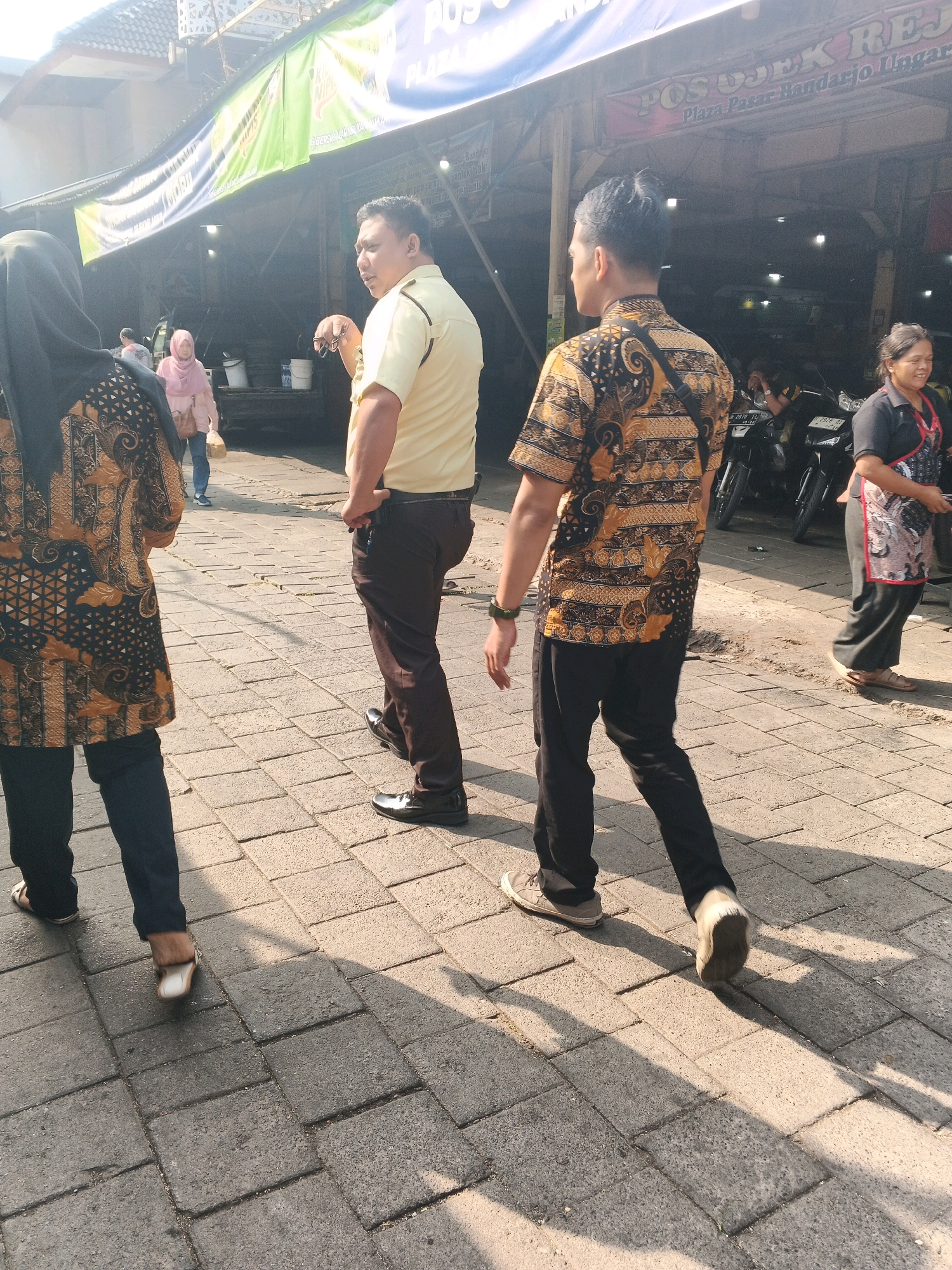 In Ungaran, Indonesia, two men and two women are captured in a daytime street scene. One man, wearing a yellow collared shirt and brown pants, stands with his left hand holding dark-rimmed glasses and his right hand on his hip. He is facing away from the camera, looking to his left. To his right, another man in a patterned batik shirt and black pants walks away from the camera, his right hand near his waist. The woman in the foreground, on the left, wears a black hijab and a patterned batik top with dark pants and is walking away from the camera. Behind the man in the yellow shirt, a woman in a pink hijab walks away from the camera carrying a bag. To the far right, a woman in a black shirt and apron walks away from the camera, holding a small object in her hand. The ground is paved with interlocking rectangular stones, and the scene is illuminated by sunlight casting shadows. In the background, there are motorcycles parked and shops with awnings, suggesting a commercial area. A banner with text, including "POS OJEK HANDARJO UNGARAN", is visible above the shops.