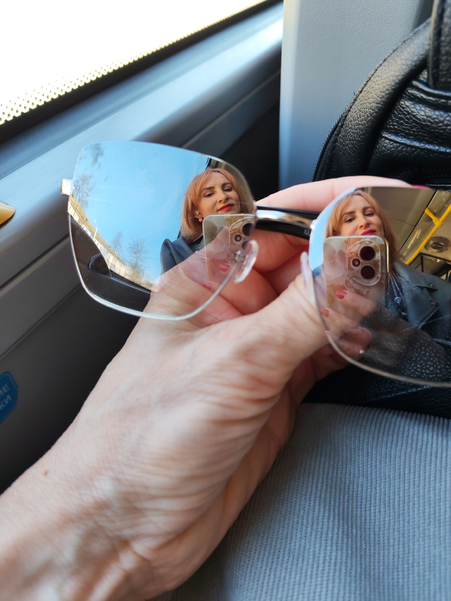 A close-up shot captures a hand holding a pair of rimless or semi-rimless sunglasses, reflecting a woman, her smartphone, and the urban environment outside a vehicle. The woman, with reddish-blonde hair and red lipstick, is visible in the lenses' reflection, looking towards the camera with a neutral expression. She is wearing a dark jacket, possibly leather. Her smartphone, with a floral-patterned case and two camera lenses, is also clearly reflected in the right lens of the sunglasses, indicating she is holding it to take the picture.
The hand holding the sunglasses is pale-skinned with visible veins and light pink fingernail polish. In the background, to the right, a black, textured bag is visible. The surrounding environment suggests the photo is taken inside a public transport vehicle, likely a bus or tram, featuring gray interior panels and a window with a white perforated trim along the top. Outside the window and vividly reflected in the left sunglass lens, bare trees, buildings, and a bright blue sky indicate a clear, sunny daytime. On the left side of the frame, a blue sticker or sign shows partial Cyrillic text, reading "МЕКИ", common in Russian public transport. The scene likely takes place in Chaikovsky, Russia.