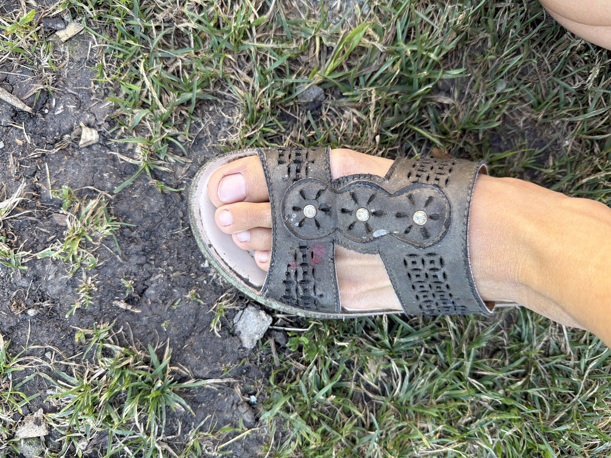 A close-up, top-down shot shows a person's left foot wearing a dark gray, open-toed sandal. The sandal has cut-out patterns and decorative circular embellishments with small, clear stones. The foot, with light skin and short, natural-colored toenails, is positioned on uneven ground covered with dry grass, dirt, and scattered small stones. The scene appears to be outdoors, possibly in a park or undeveloped area, consistent with the outskirts of a rural settlement like Petryaksa, Russia, where such terrain is common.
