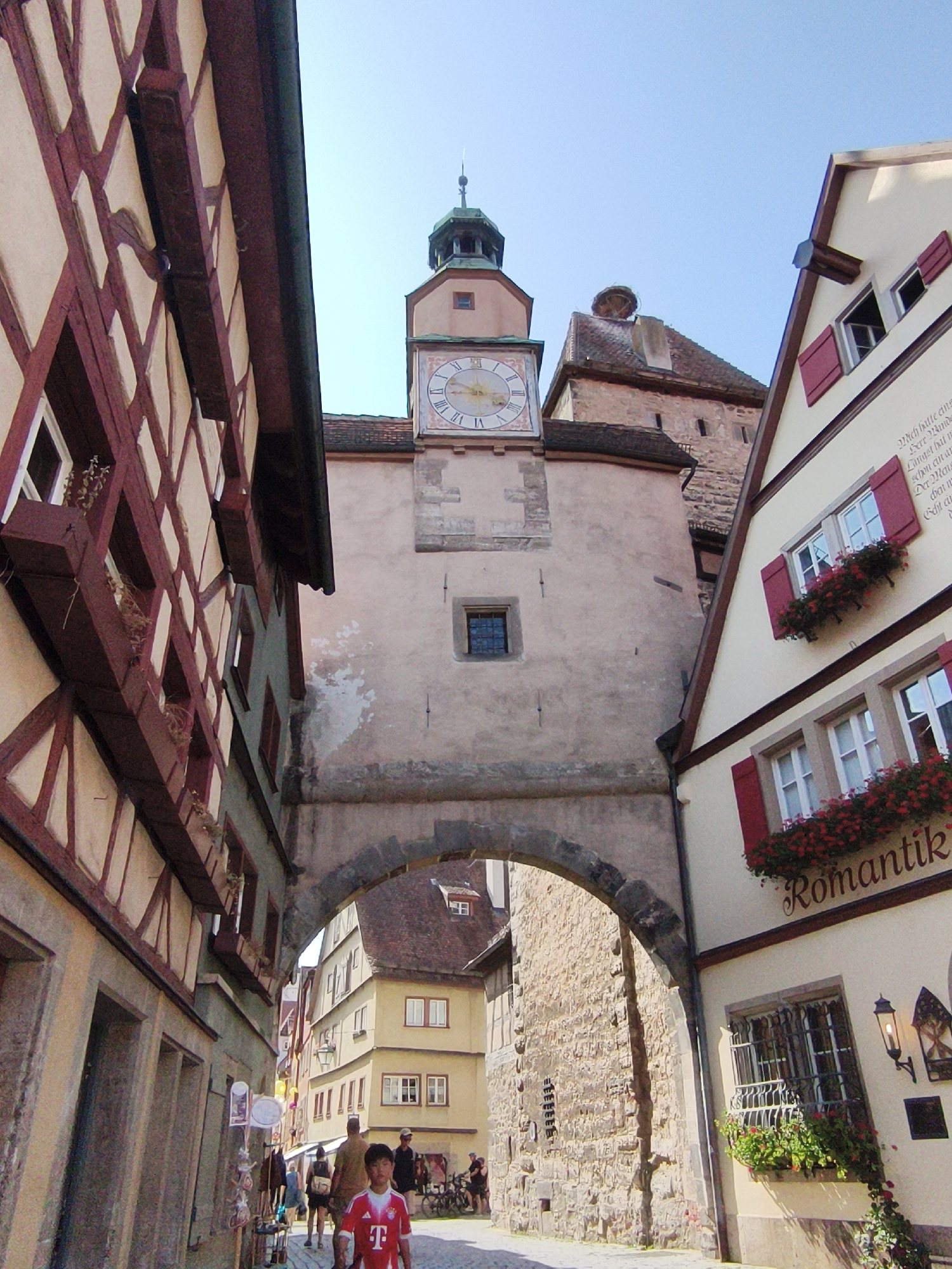 The image depicts a view through a stone archway onto a narrow street in Rothenburg ob der Tauber, Germany. The architecture is characterized by half-timbered buildings with distinctive dark wooden beams and cream-colored plaster walls. A prominent clock tower with a Roman numeral clock face is visible in the background, centrally located within the archway. On the right side of the street, a building features a sign with the word "Romantik" in cursive script and red shutters on its windows, adorned with flower boxes overflowing with red flowers. Several people are visible in the scene, including a young boy in a red t-shirt walking towards the viewer in the foreground. Other individuals are further down the street, some appearing to be shoppers or tourists. The sky is clear and blue, indicating fair weather. The overall impression is of a historic European town with well-preserved medieval architecture.